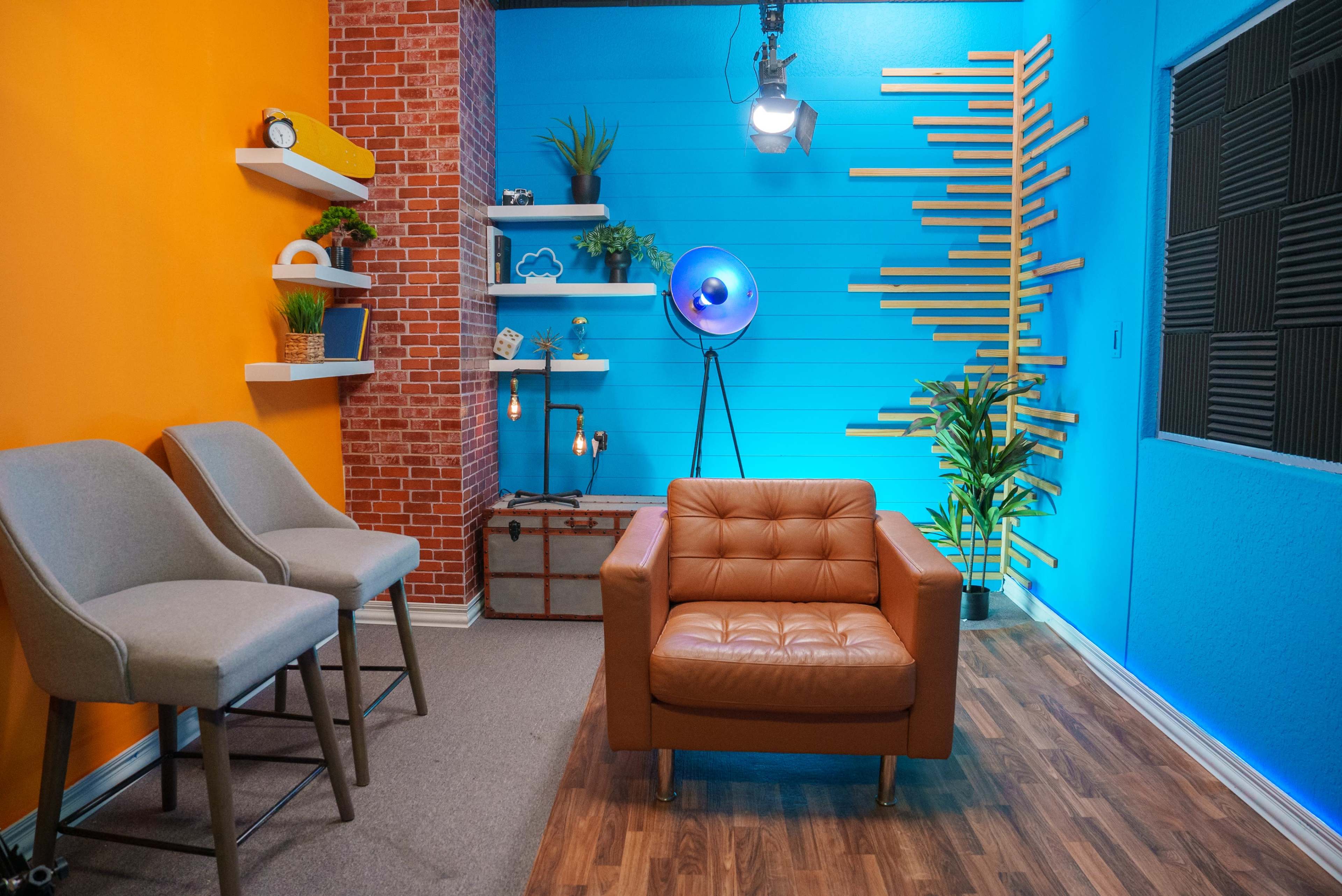 The image shows a modern studio setup with an orange and blue painted wall, stylish furniture, and decorative shelves featuring plants and lighting.