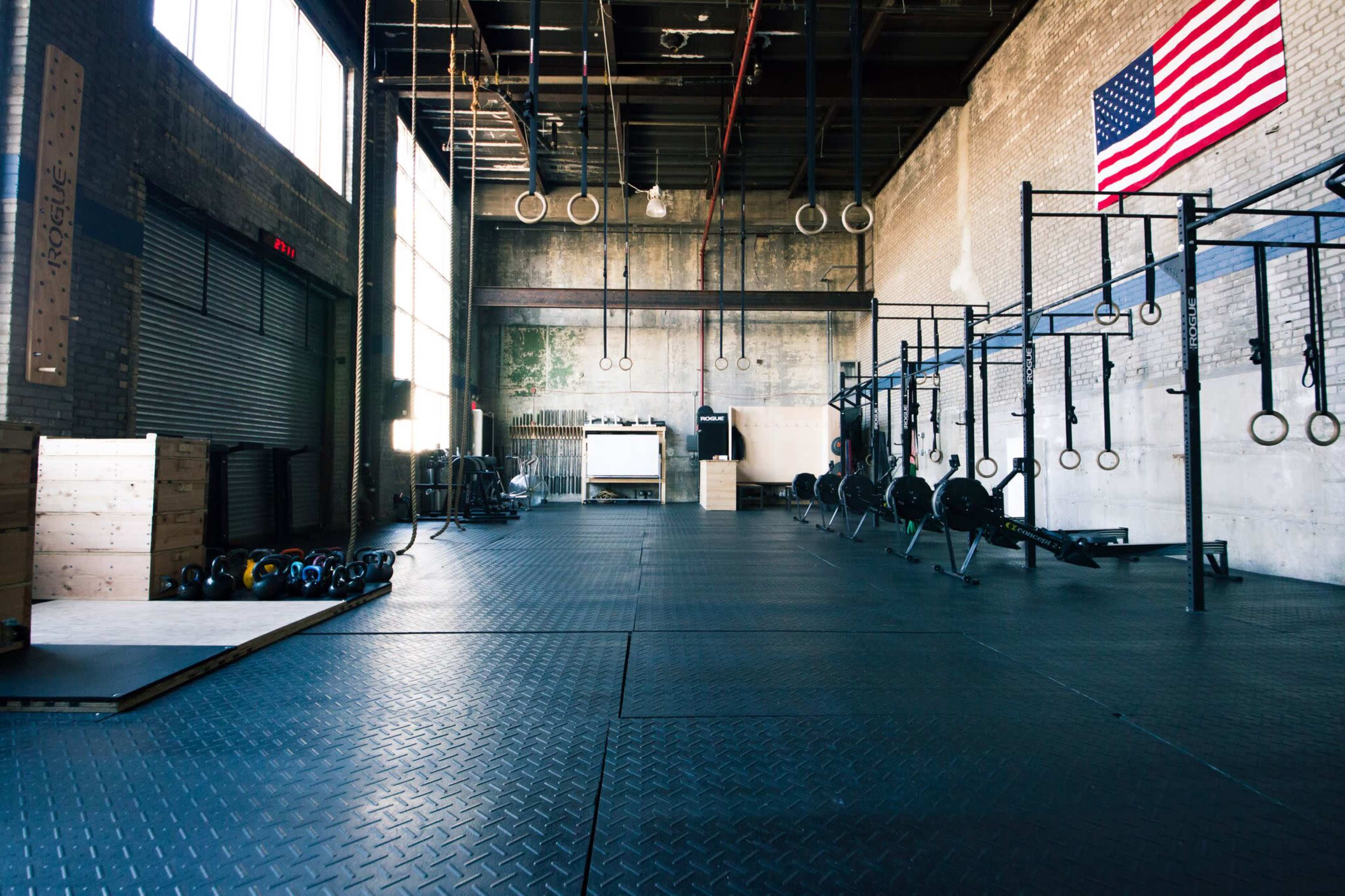 The image shows an interior of a gym with various equipment, including weights and gymnastic rings, along with an American flag hanging on the wall.