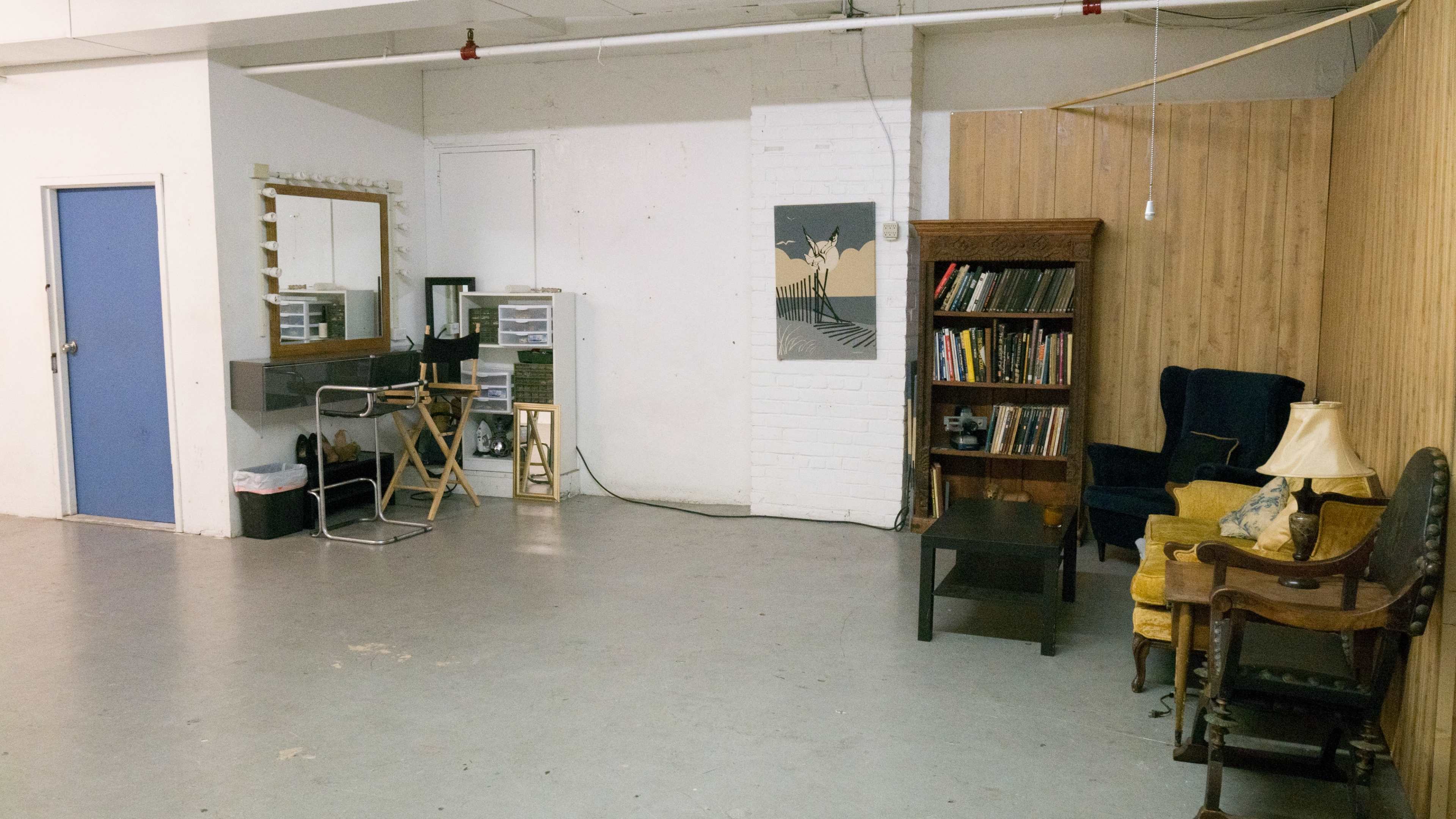 A spacious room with a makeup station, a bookshelf filled with books, and various seating options against a plain backdrop.