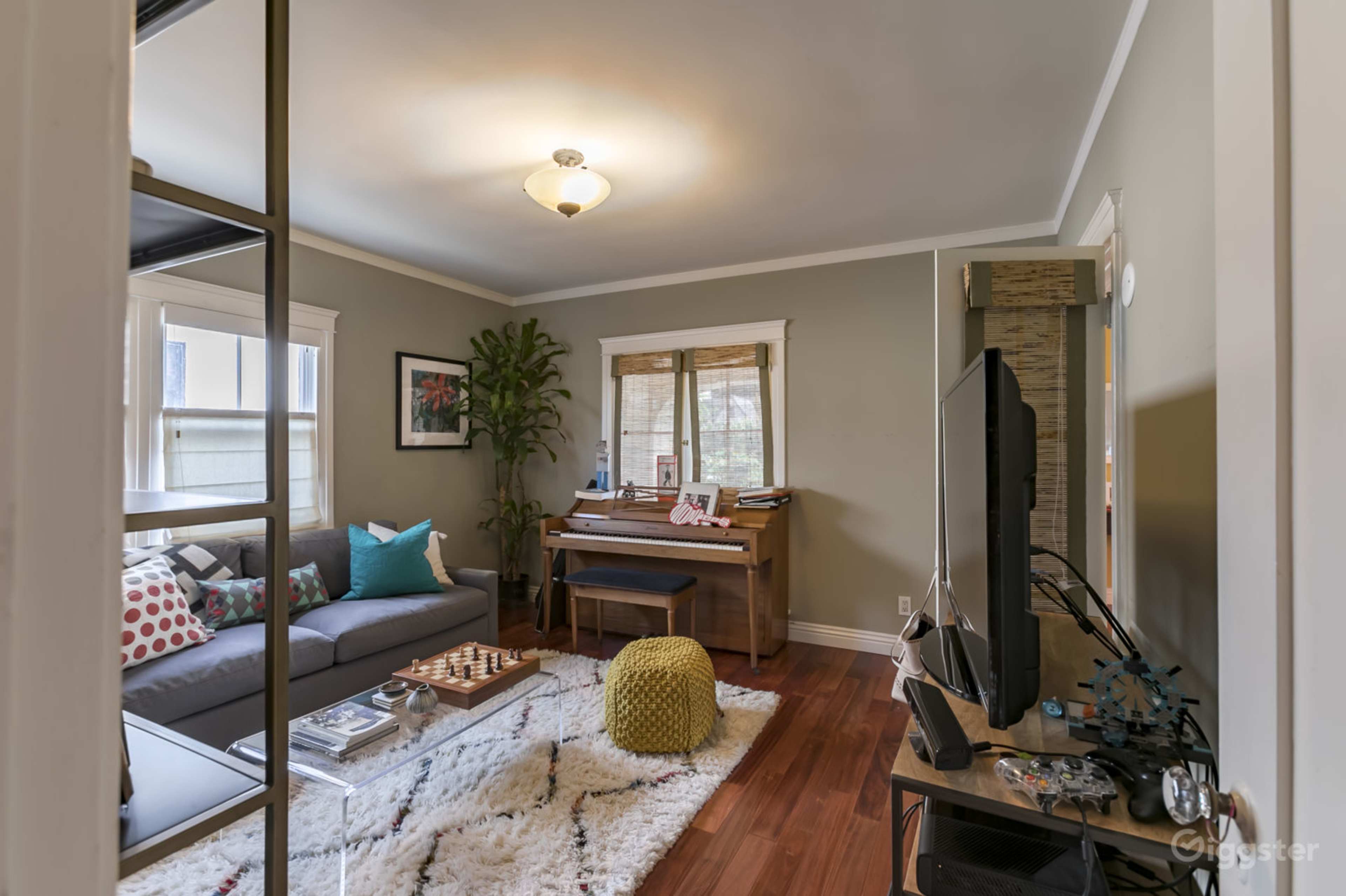 A cozy living room features a gray sofa, a piano, a coffee table, and a large area rug, with natural light coming through the windows.