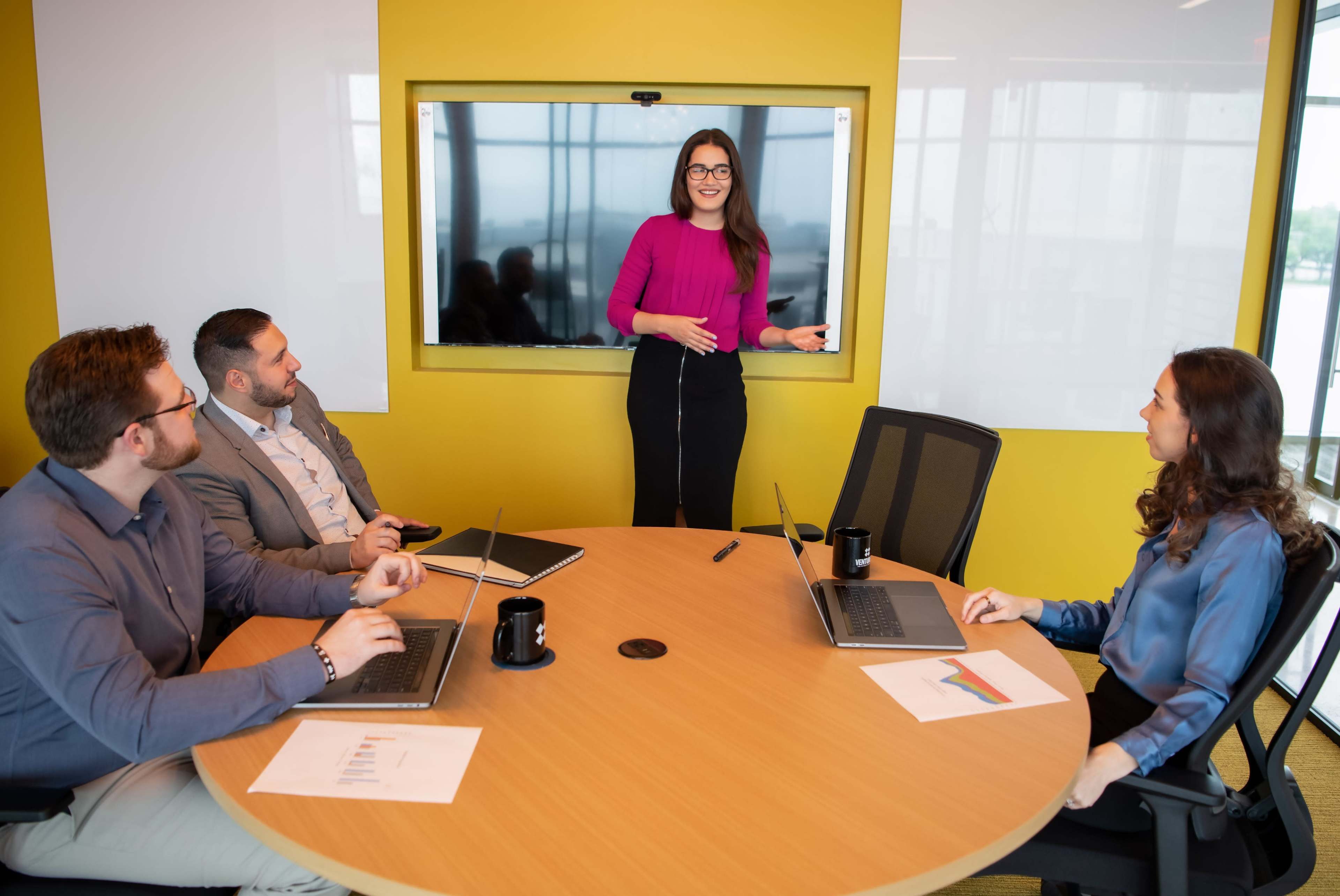 A group of four professionals are seated at a round table in a conference room, with one woman standing and presenting in front of a large screen.