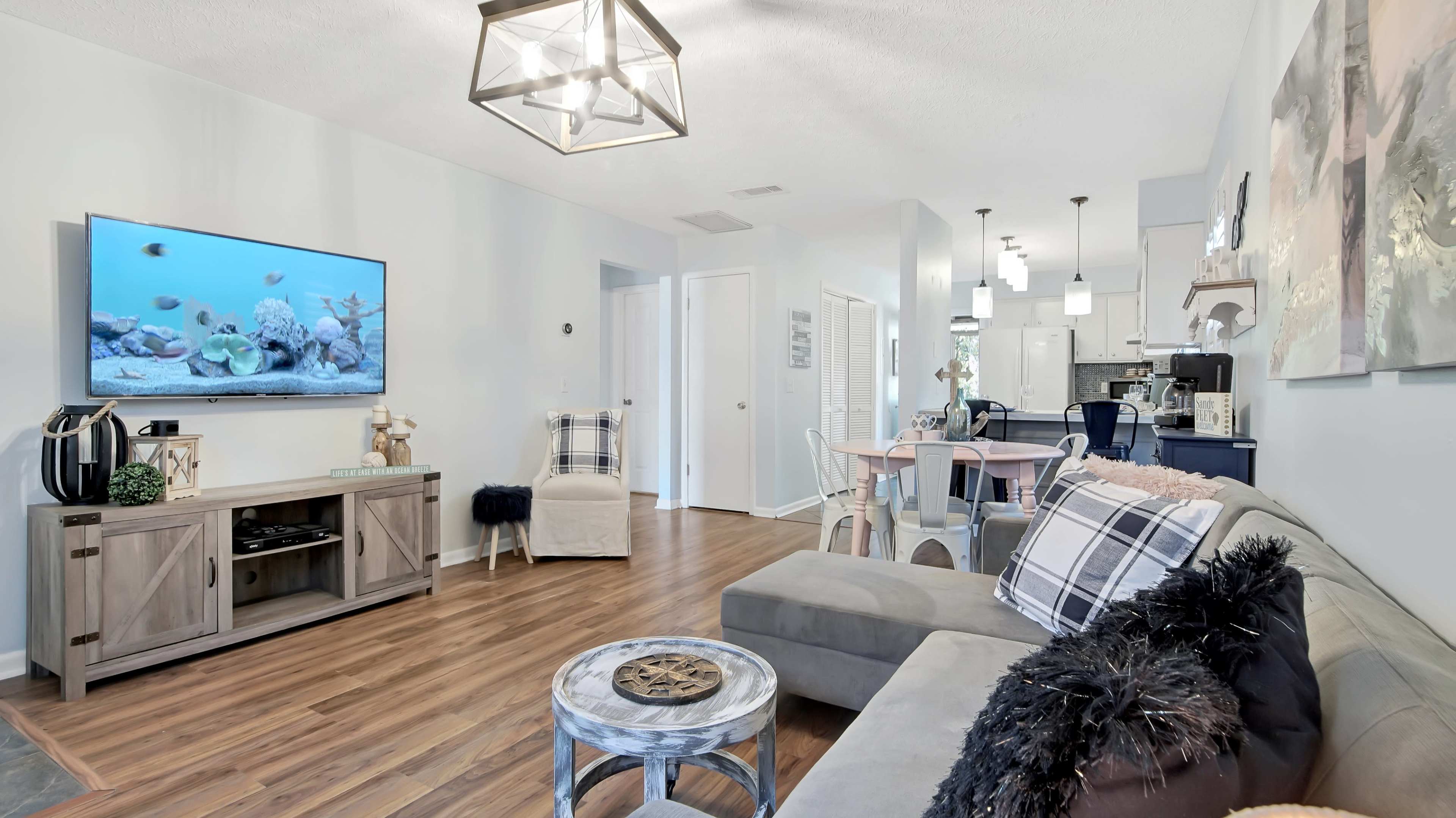 A spacious living room with a television mounted on the wall, a sofa with decorative pillows, a coffee table, and an adjoining dining area with a light-colored table and chairs.