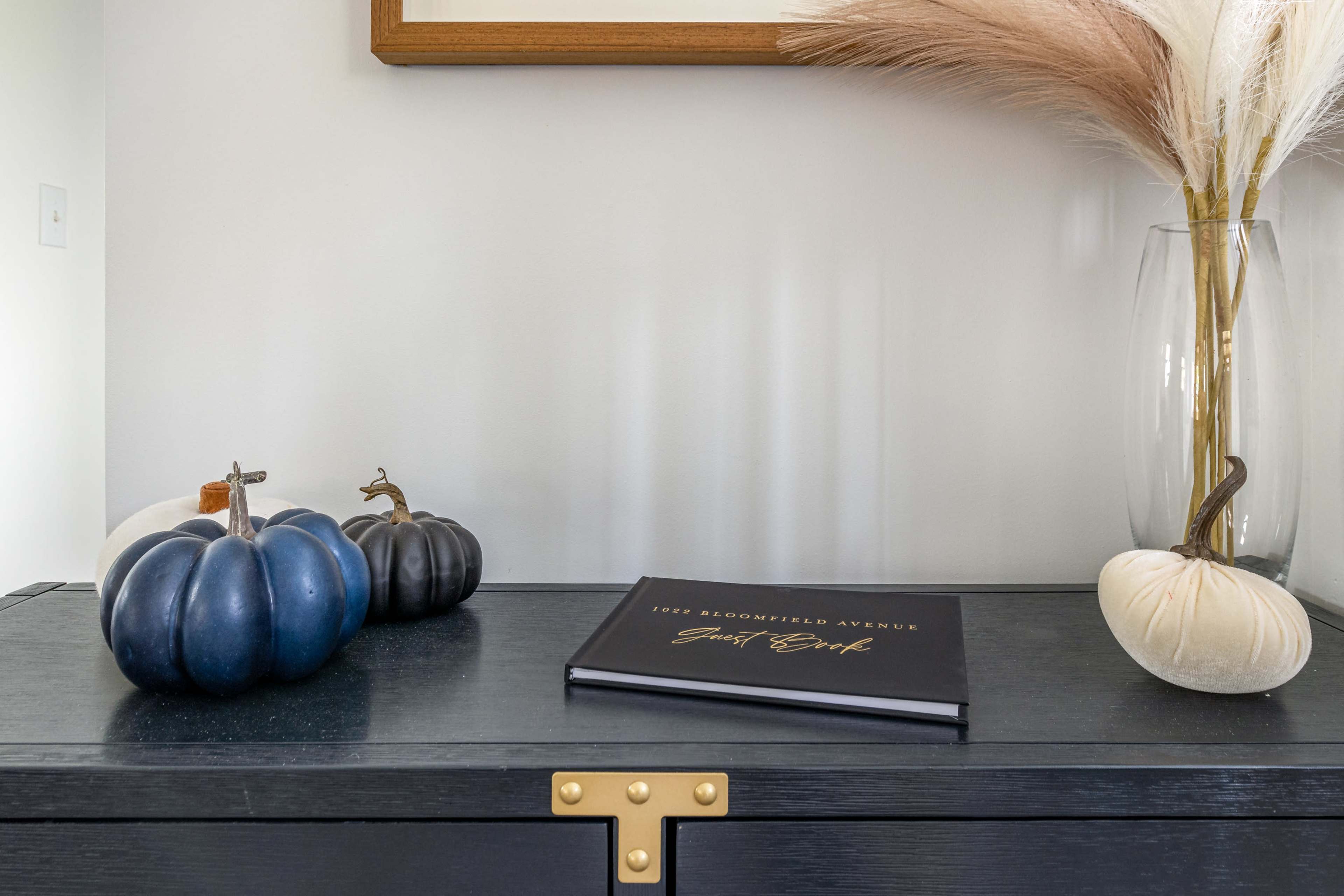 A black cabinet displays decorative pumpkins and a closed book titled "Guest Book" alongside a vase with dried grass.