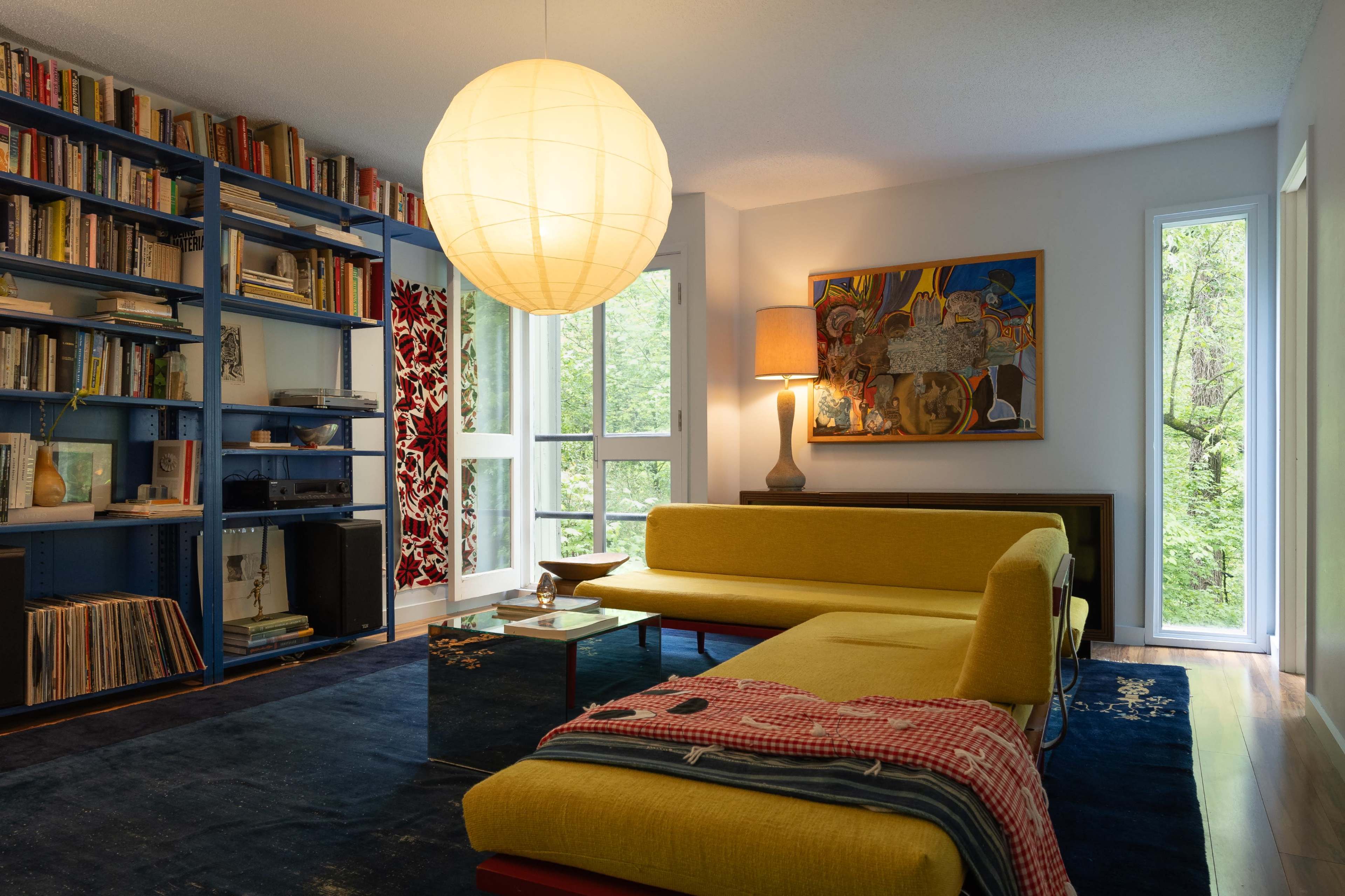 A modern living room featuring a yellow couch, a large round pendant light, bookshelves filled with books, and windows overlooking greenery.