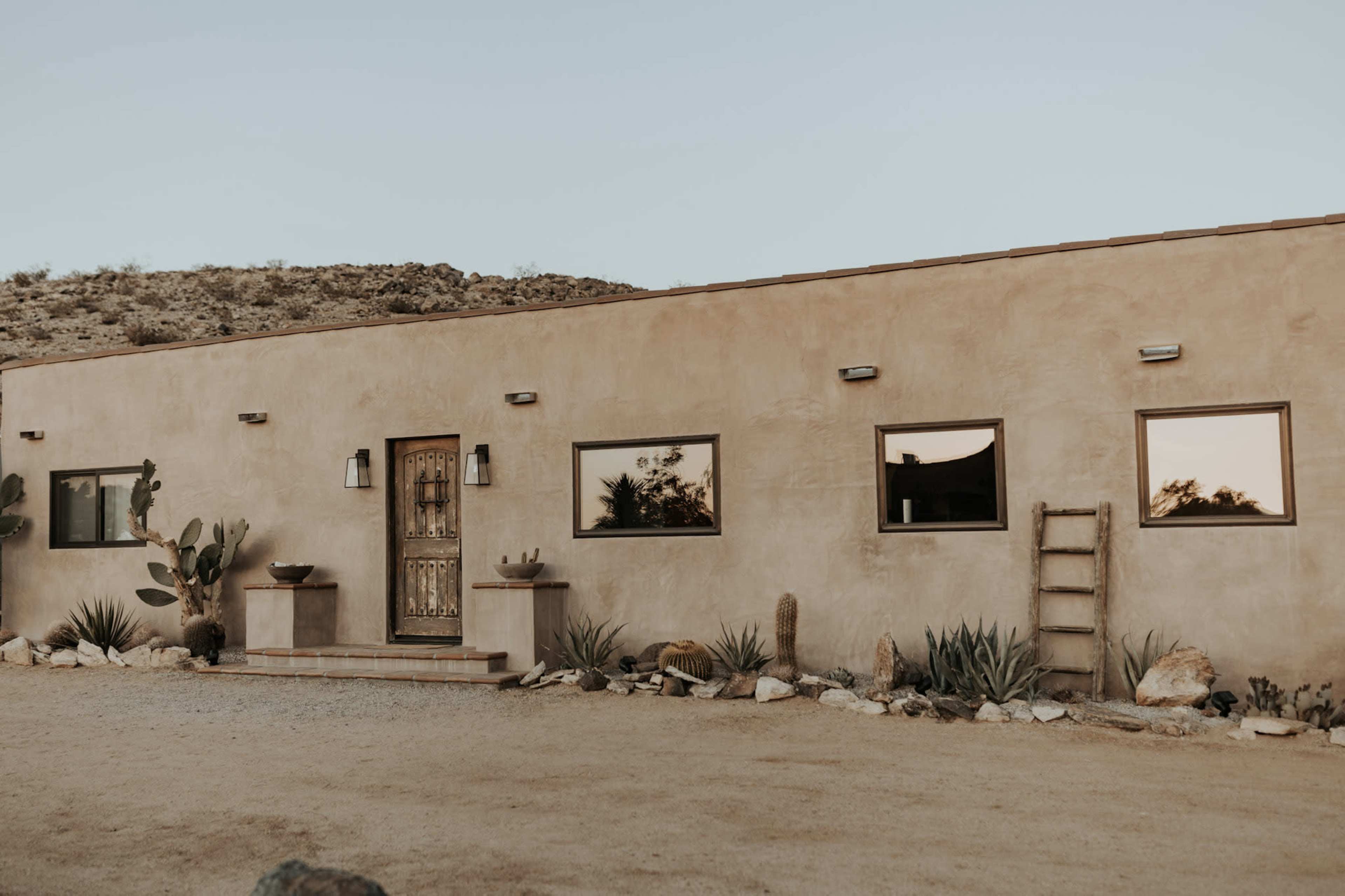 A single-story adobe-style house with a wooden front door and three windows is set in a desert landscape with gravel and various cacti.