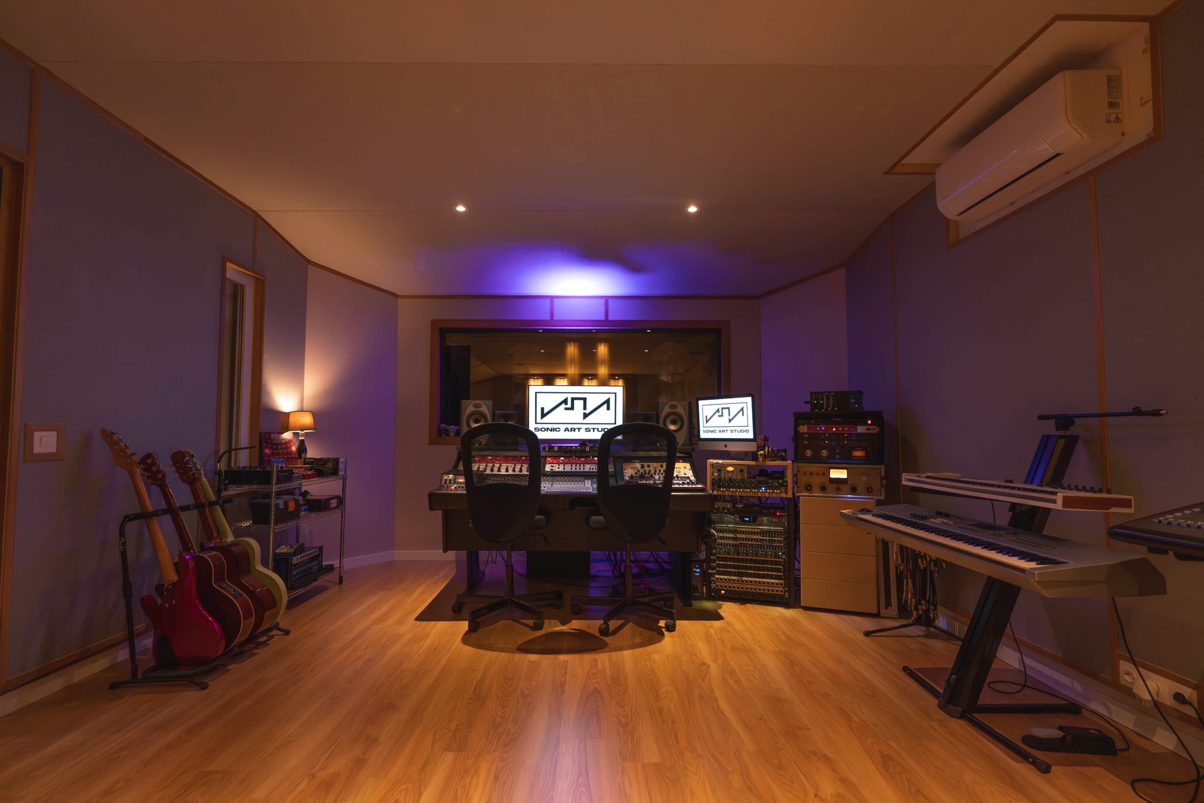 The image shows a music recording studio with a central mixing console, two computer monitors, and various musical instruments, including electric guitars and keyboards, arranged around the space.