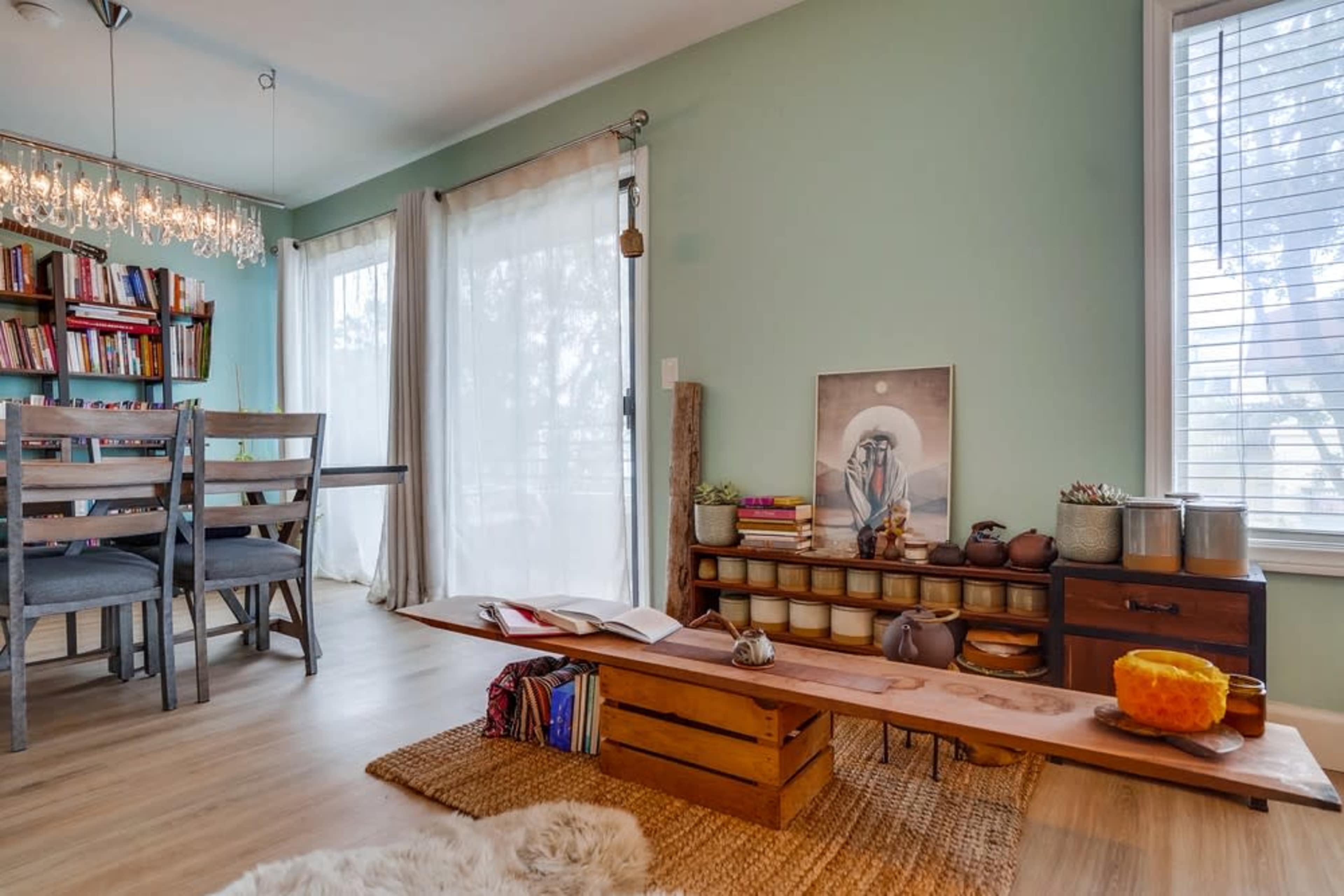A living space with a wooden bench, bookshelves, and a dining area featuring a large window with sheer curtains.