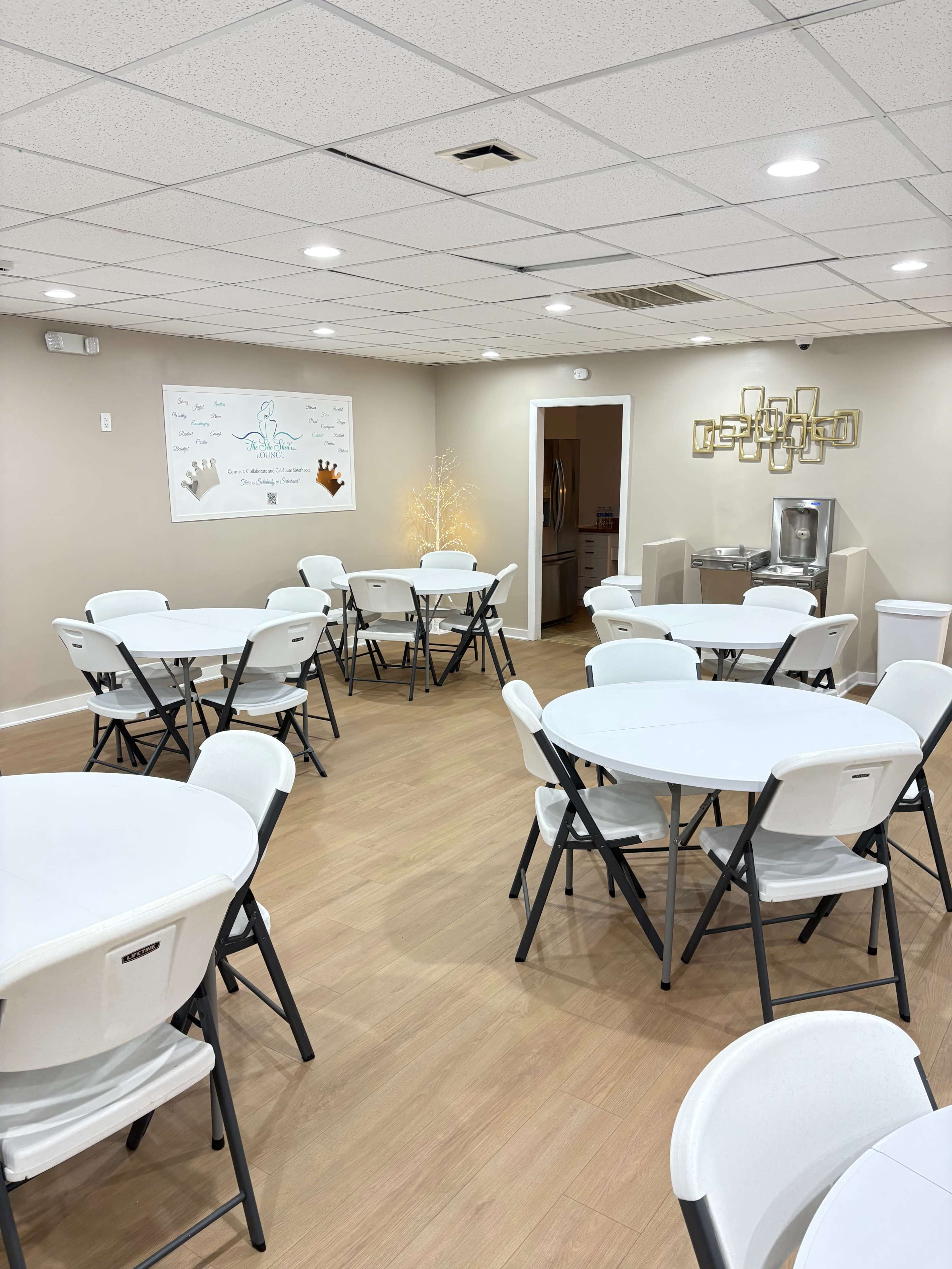 The image shows a well-lit community room with several round tables and chairs arranged neatly, a wall with decorative elements, and a water cooler in the corner.