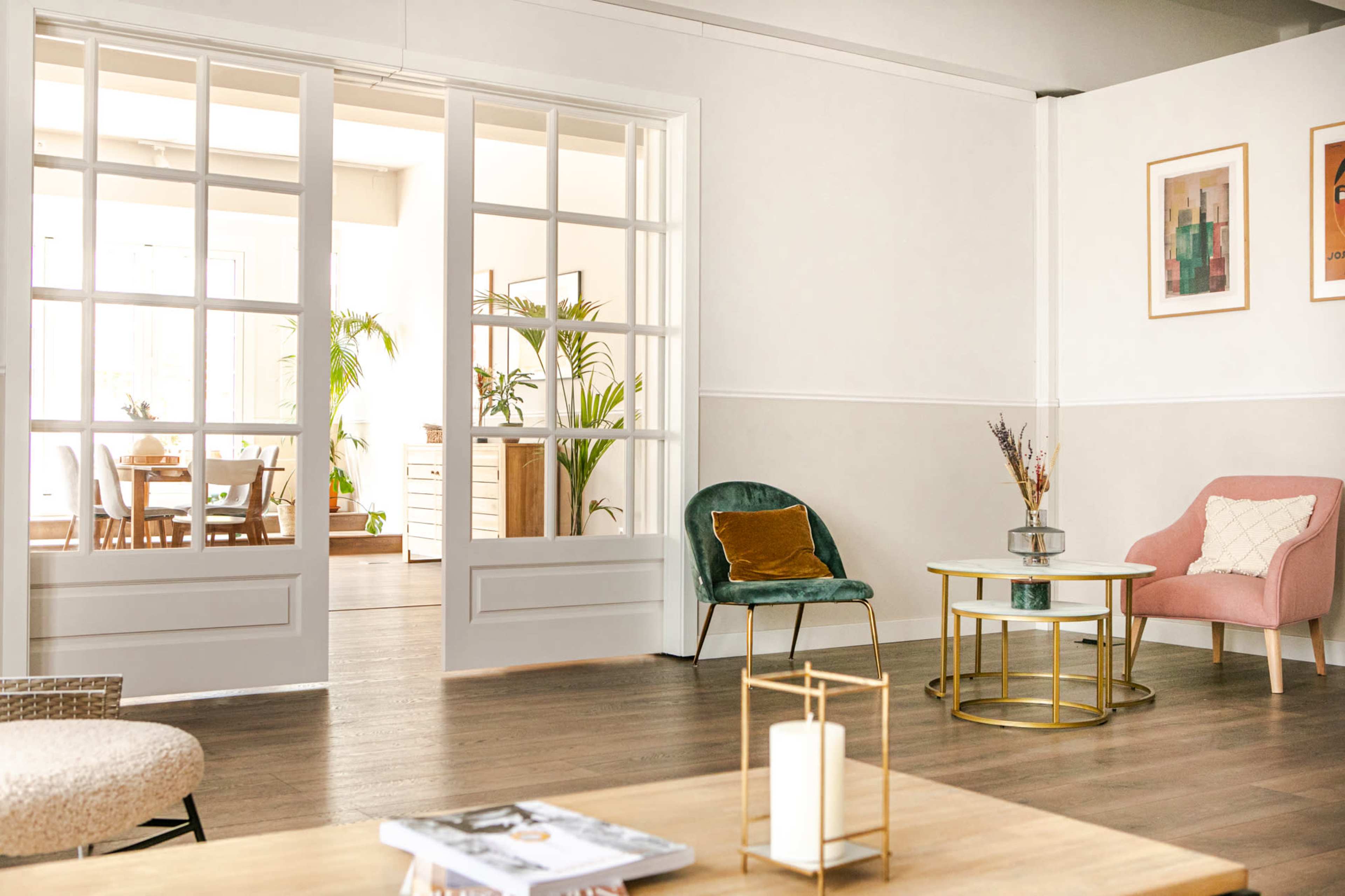 A well-lit room with glass-paneled double doors leading to another space, featuring modern furniture, including chairs and a coffee table, surrounded by greenery.
