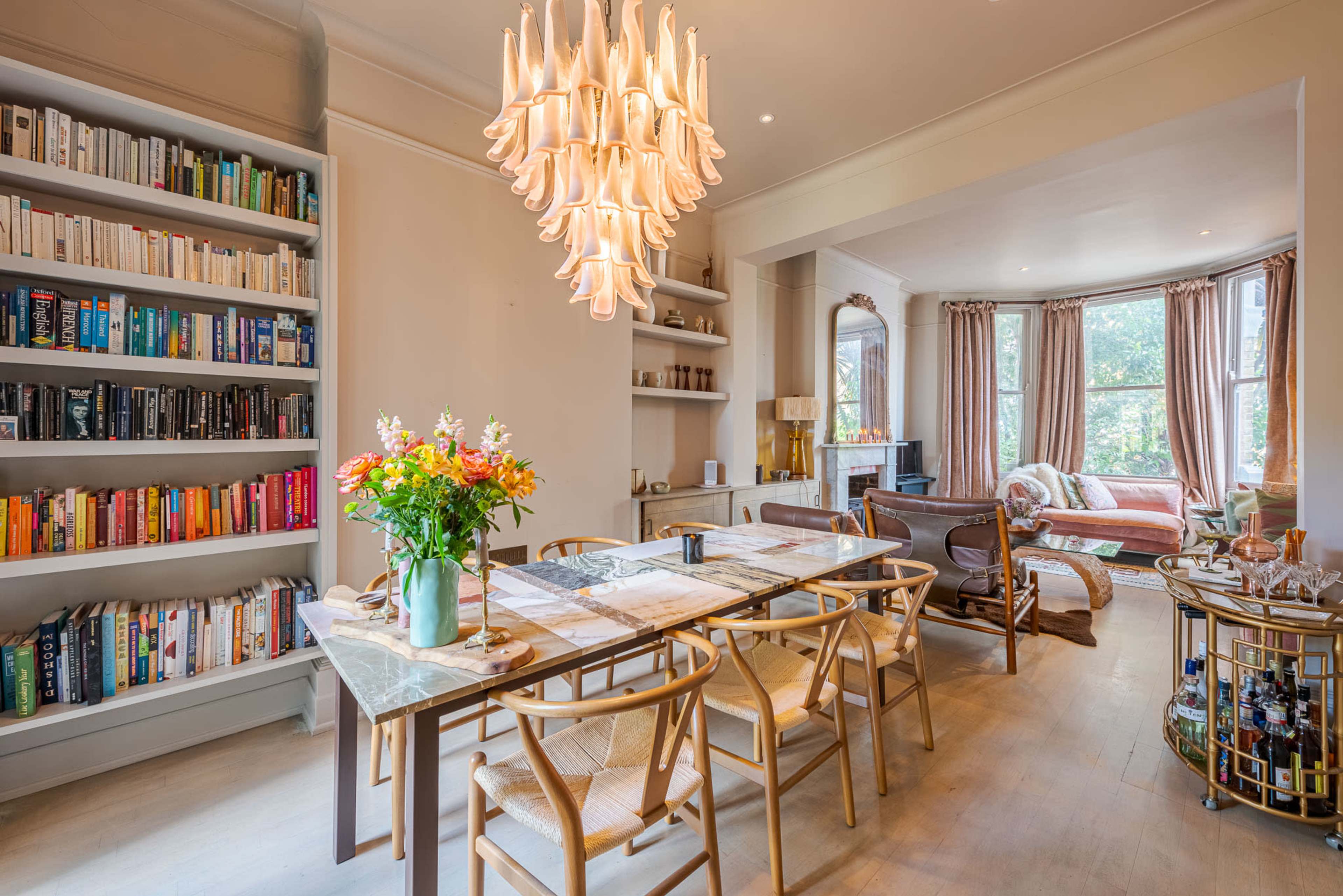 A well-lit dining area features a large table surrounded by chairs, a bookshelf filled with books, and a chandelier hanging overhead.