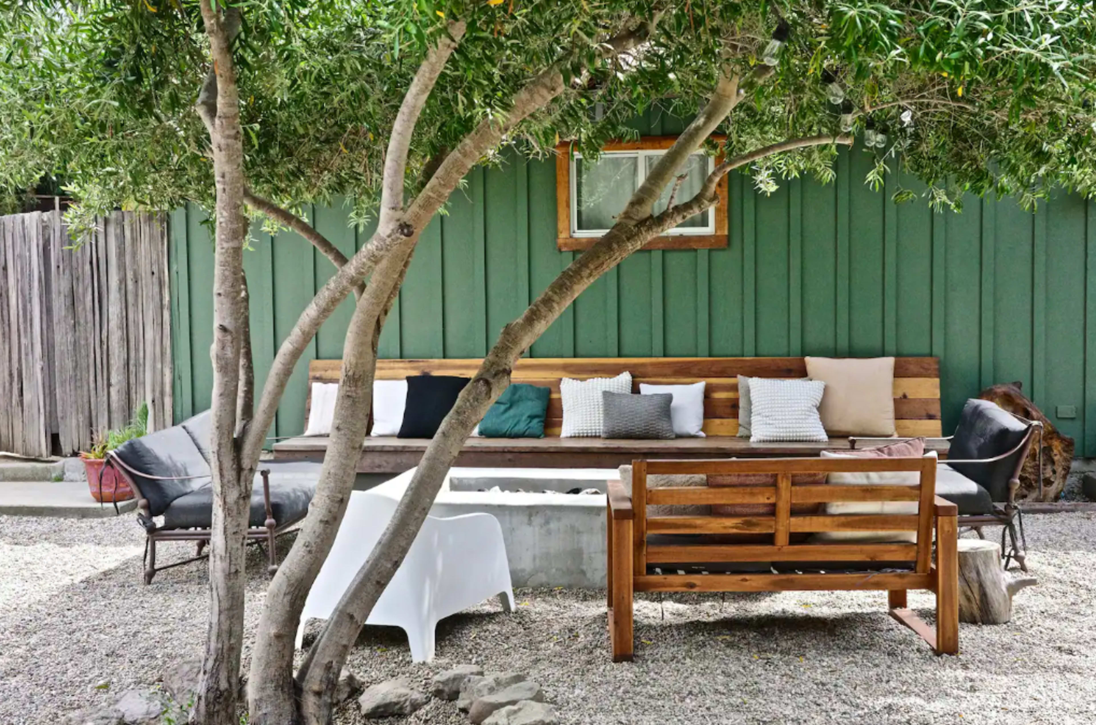 The image shows a landscaped outdoor seating area featuring a wooden bench, several cushions, and a central table under a tree, surrounded by gravel.