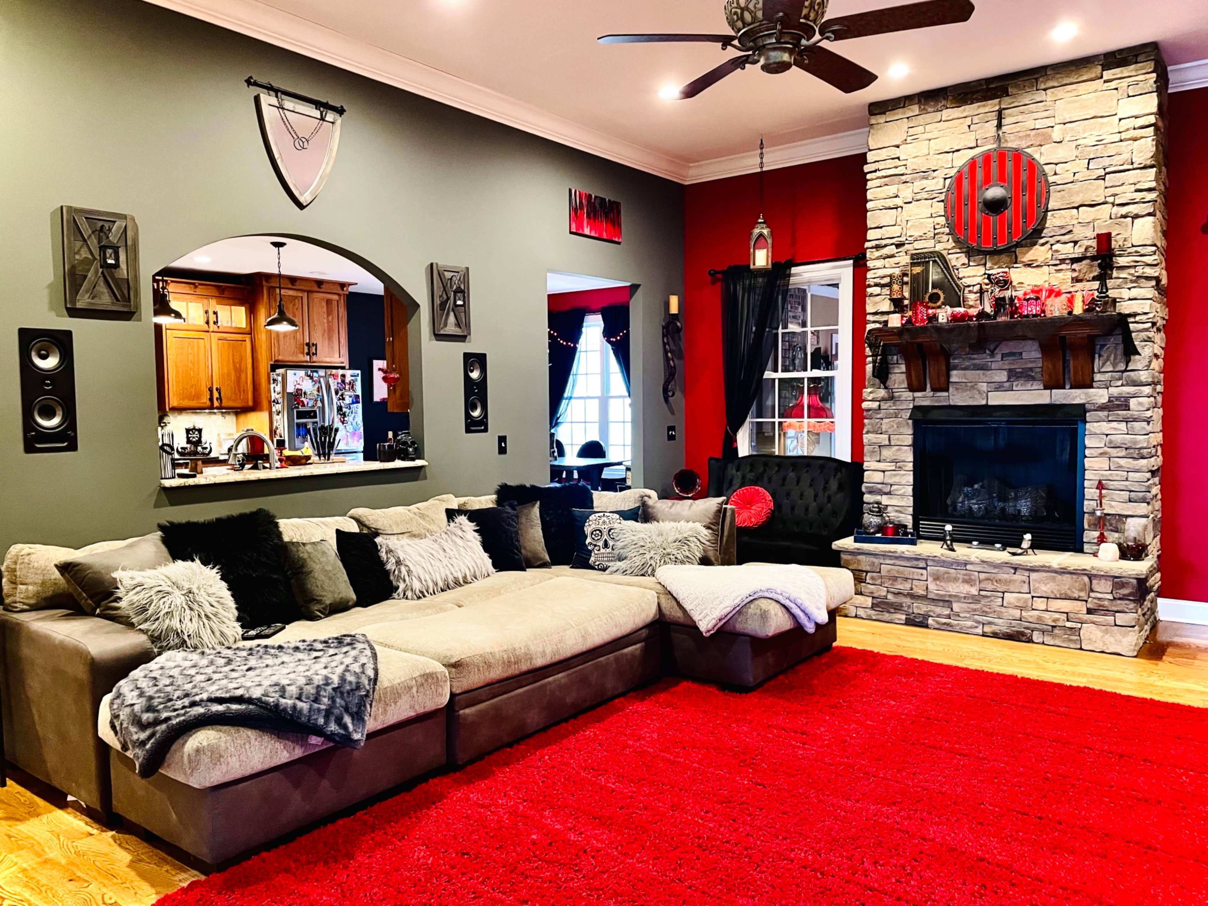 The room features a sectional sofa on a red rug, with a stone fireplace as a focal point and a kitchen visible through an archway.