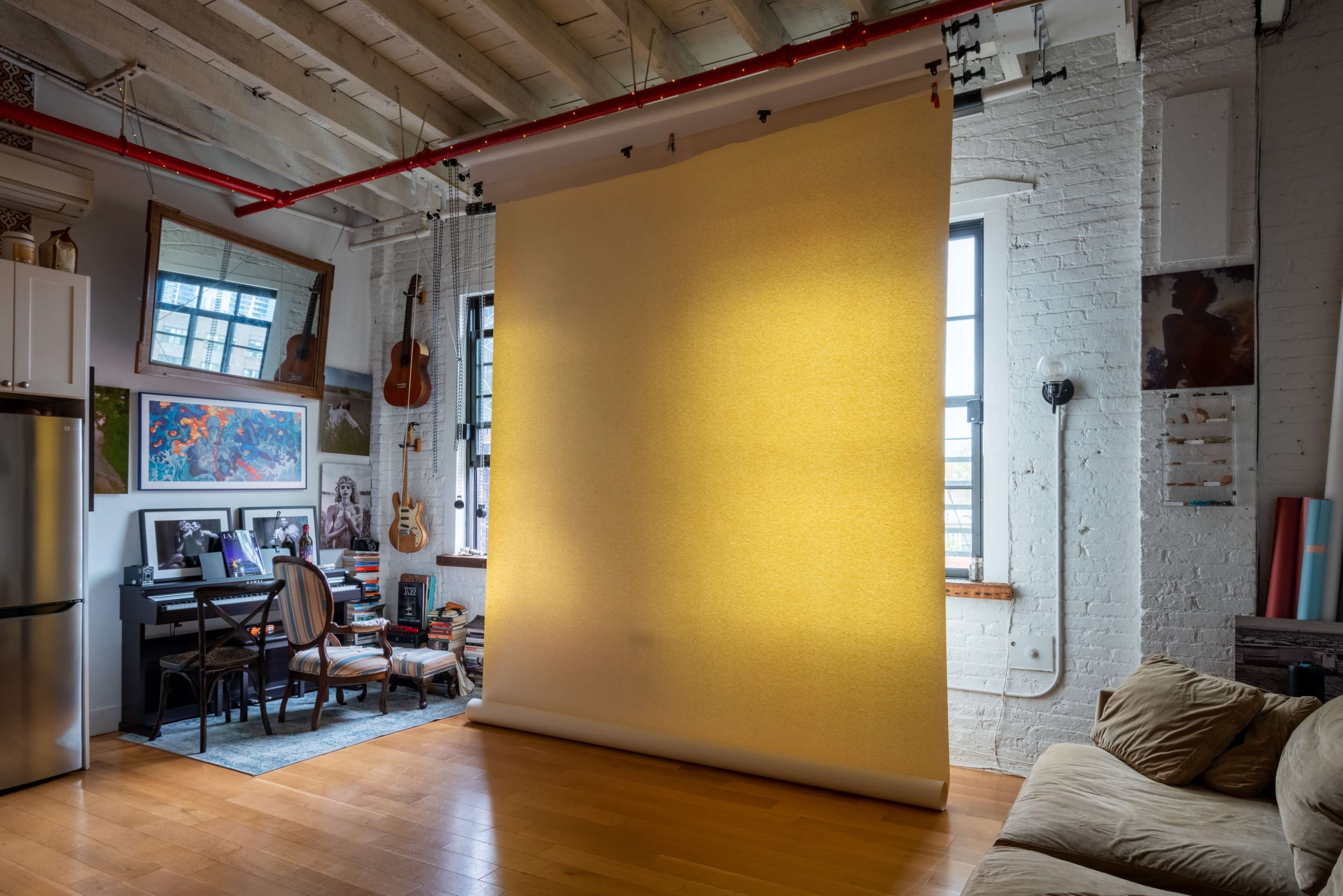 A bright yellow backdrop is set against a brick wall in a spacious room with wooden floors and various artworks displayed around.