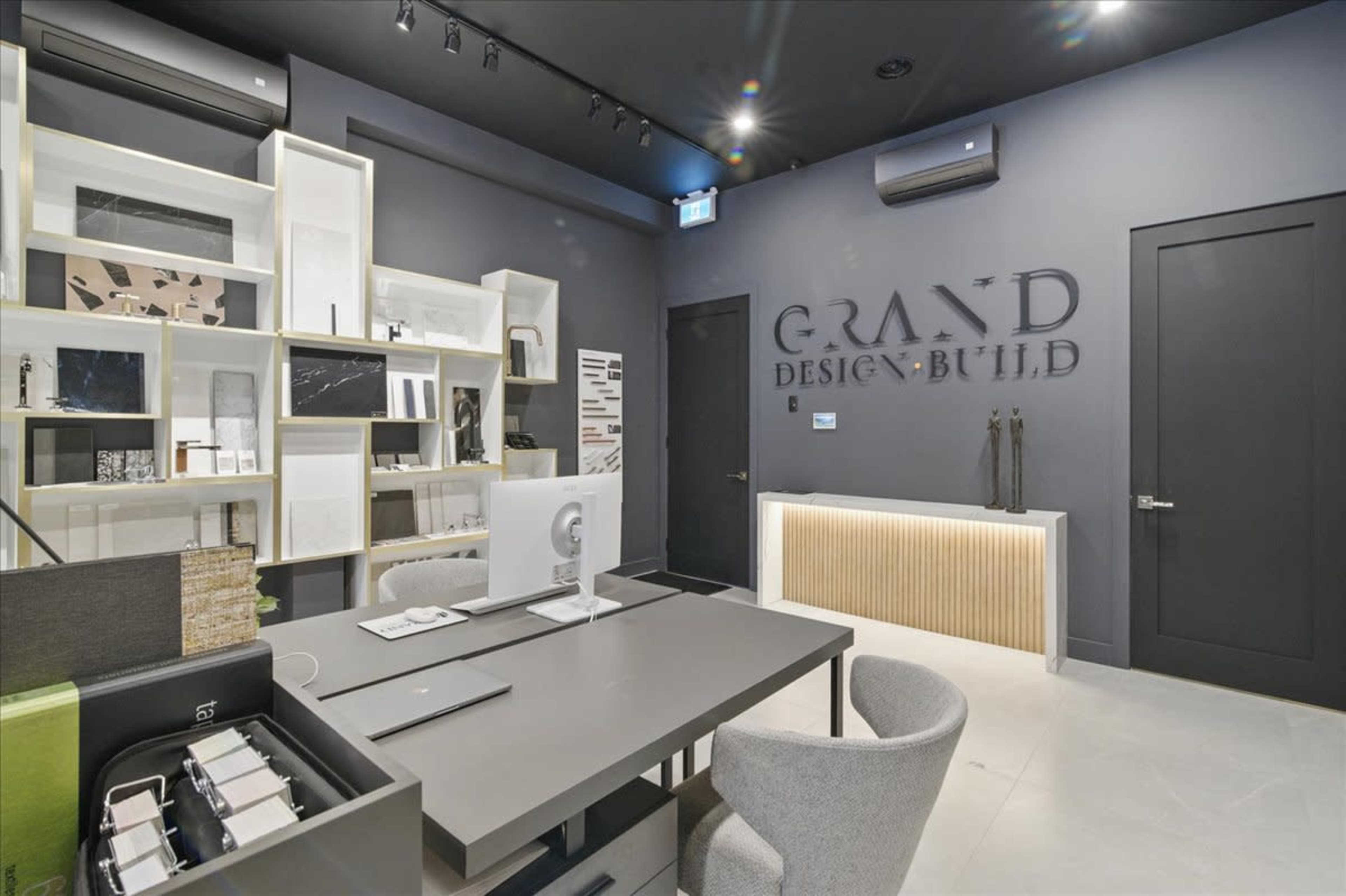 The image shows a modern office space with a gray color scheme, featuring a desk with a computer, shelves displaying design materials, and a wall with the words "GRAND DESIGN BUILD" prominently displayed.