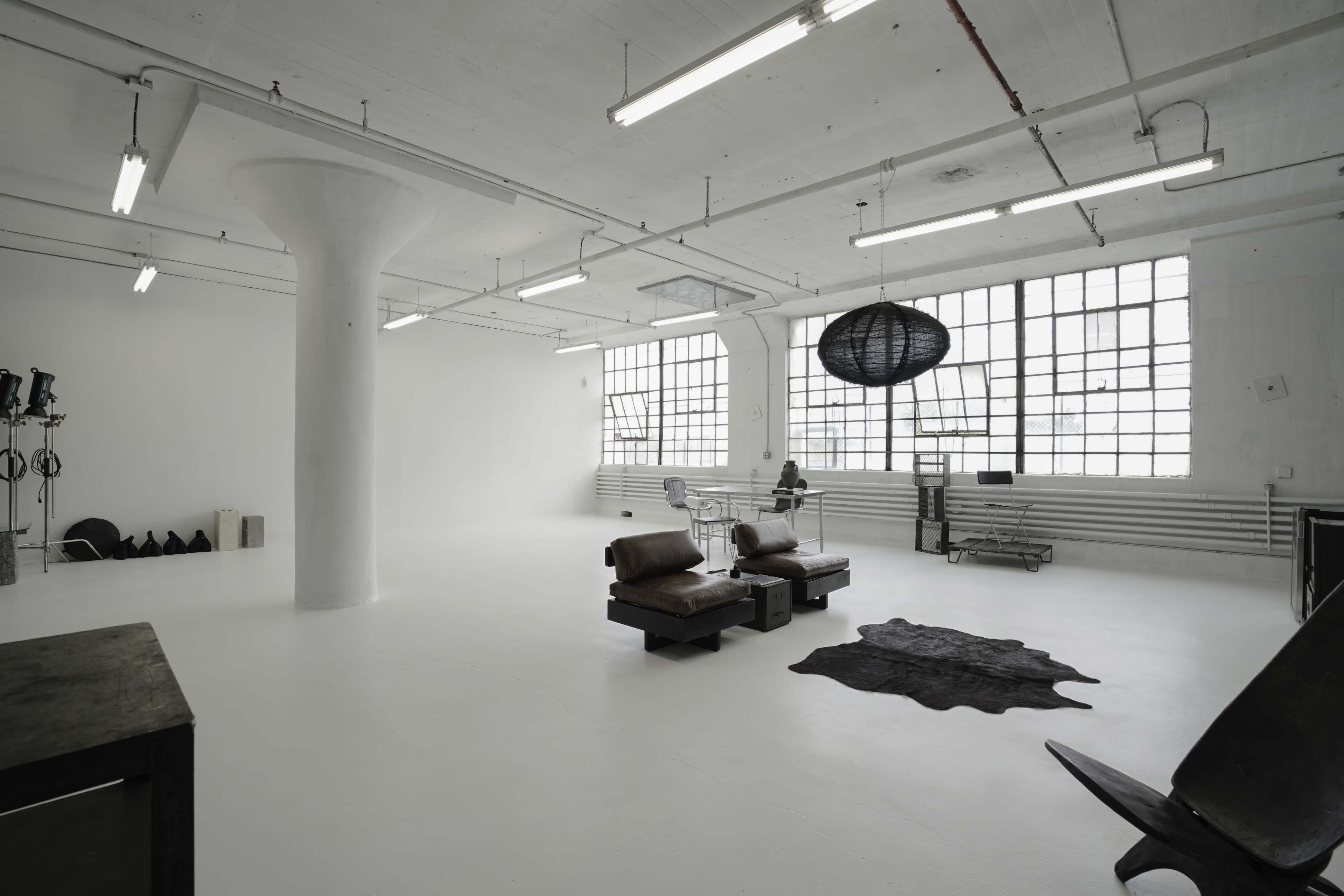 A spacious, minimally furnished room with white walls, large windows, and a few pieces of modern furniture, including two leather chairs and a round pendant light.