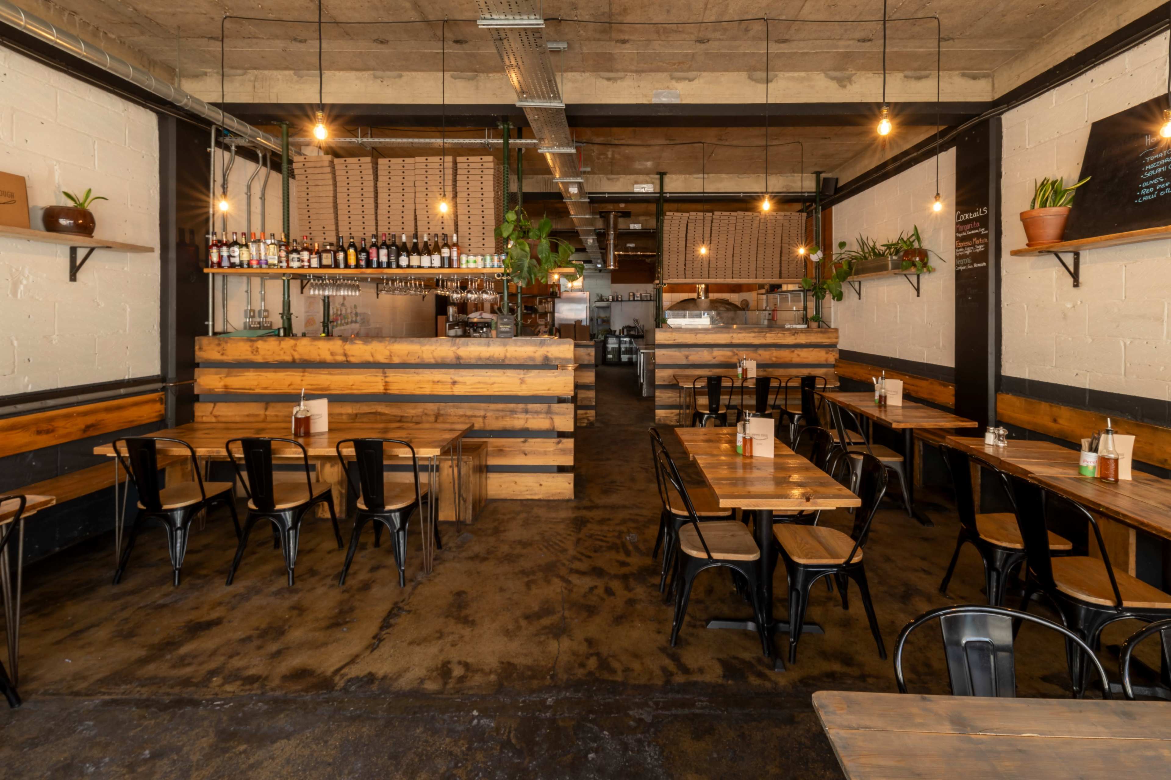 The image shows a spacious restaurant interior with wooden tables, metal chairs, and plants arranged on shelves and walls.