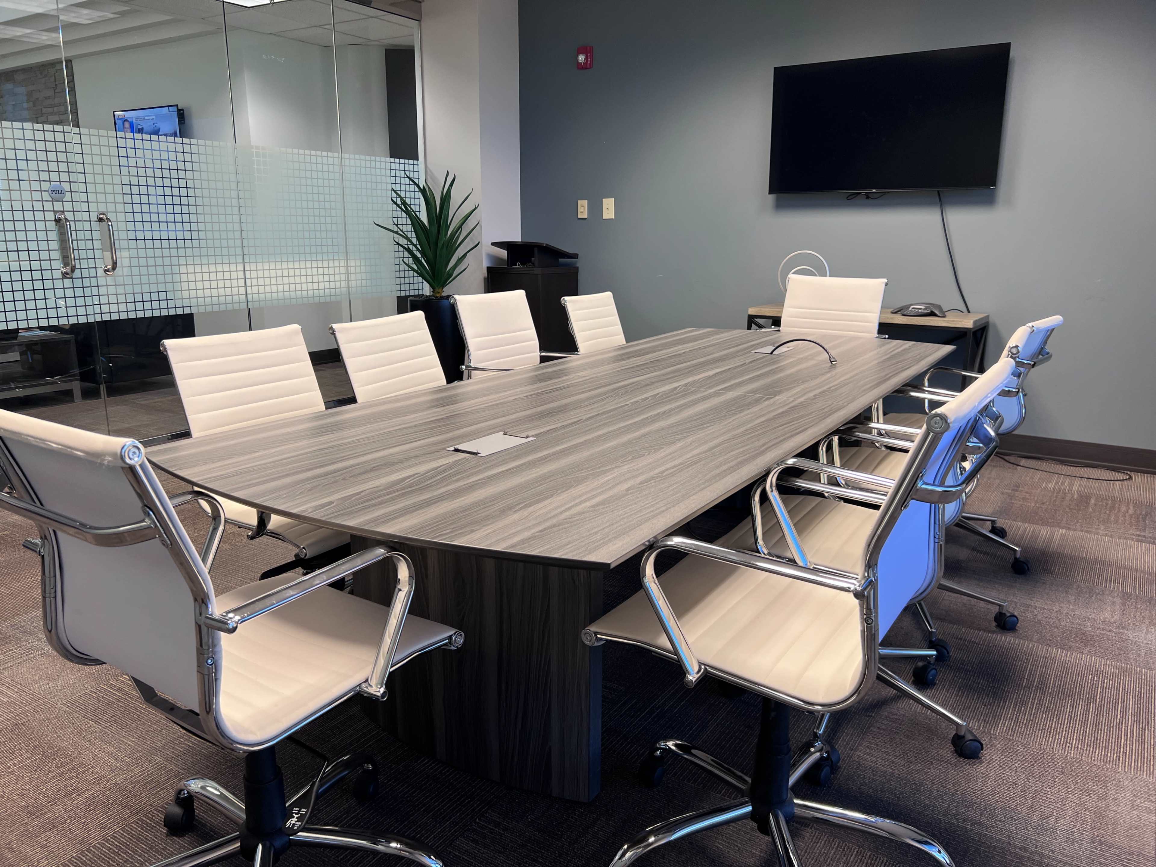 A modern conference room with a large oval table surrounded by several white leather chairs and a wall-mounted television.