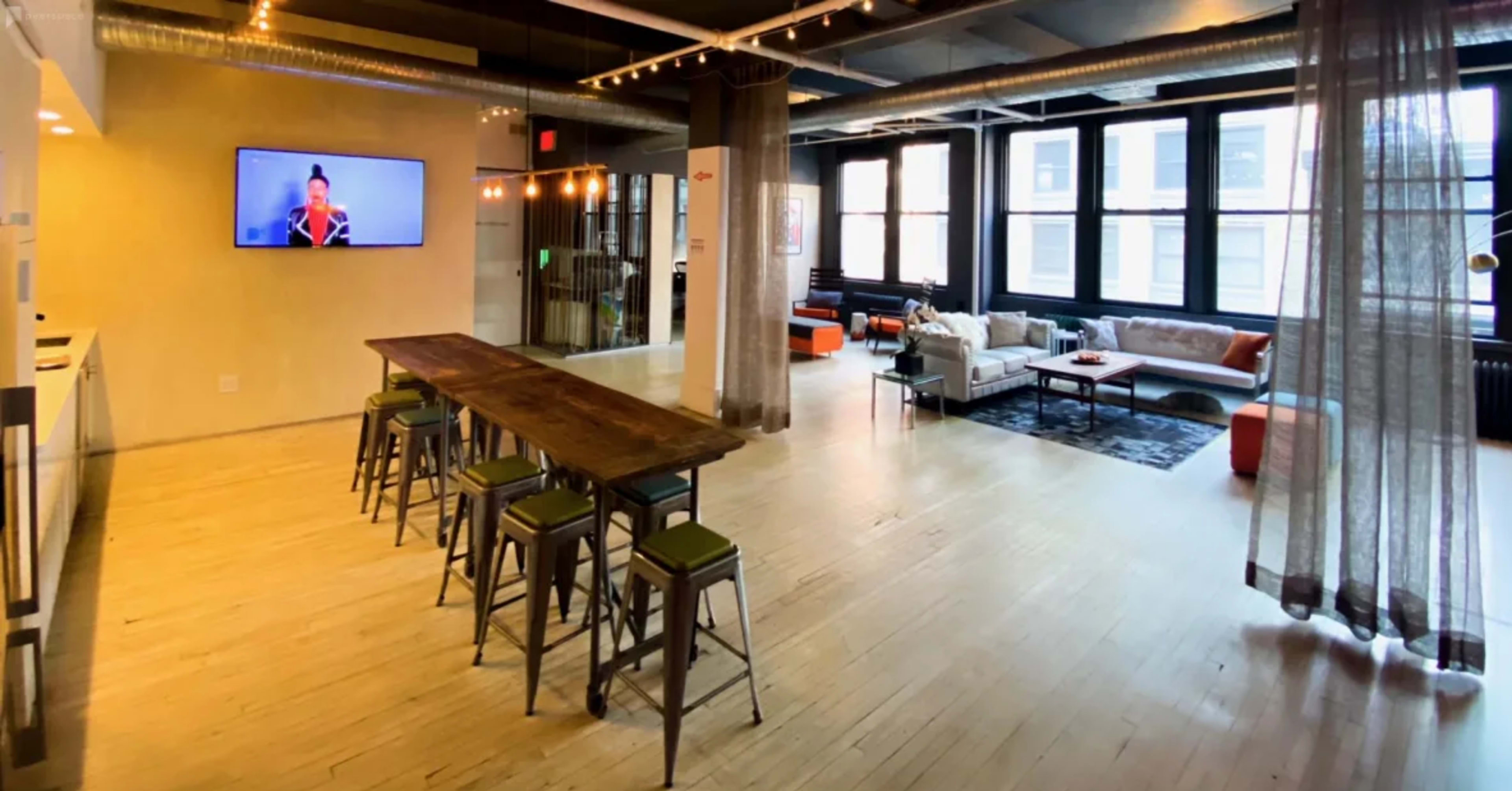 A spacious, modern indoor area with a long wooden table and metal stools, a television mounted on the wall, and a seating area with a couch, coffee table, and lounge chairs.