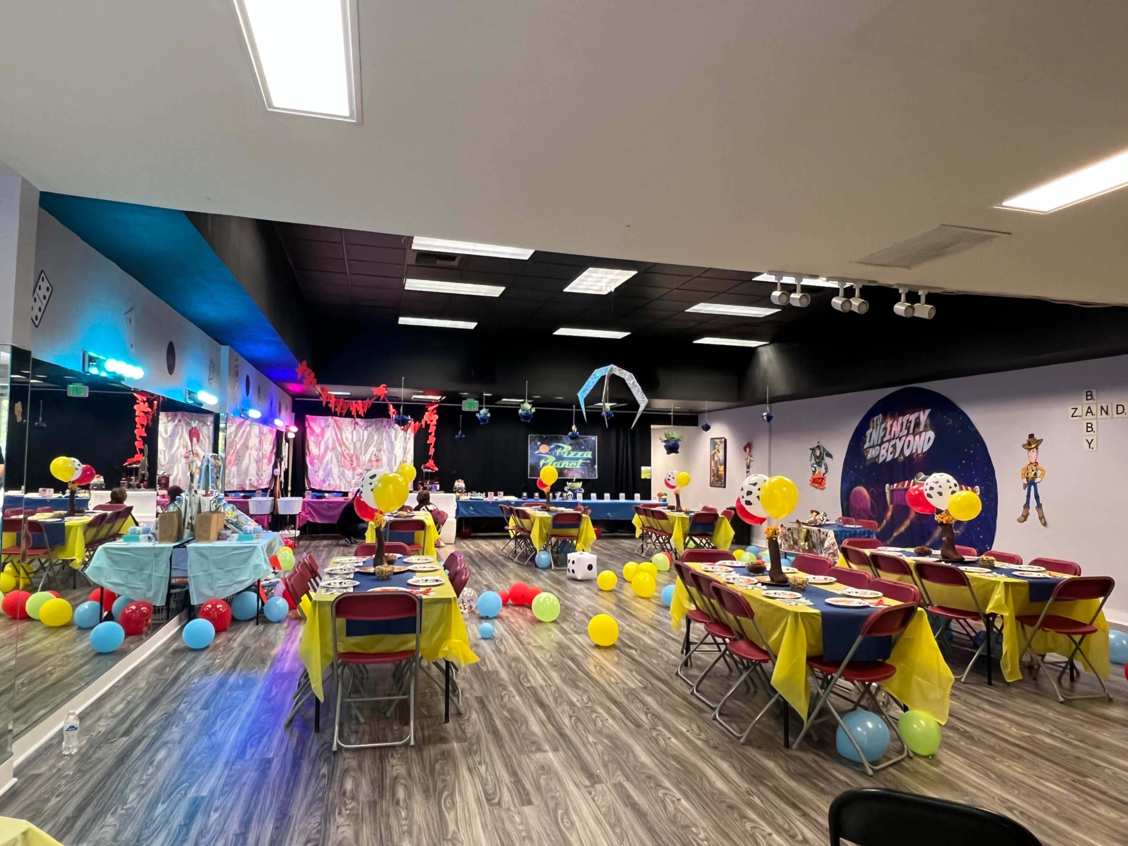 A brightly decorated party venue with tables set for a celebration, featuring balloons, colorful tablecloths, and a stage area with a projected screen.