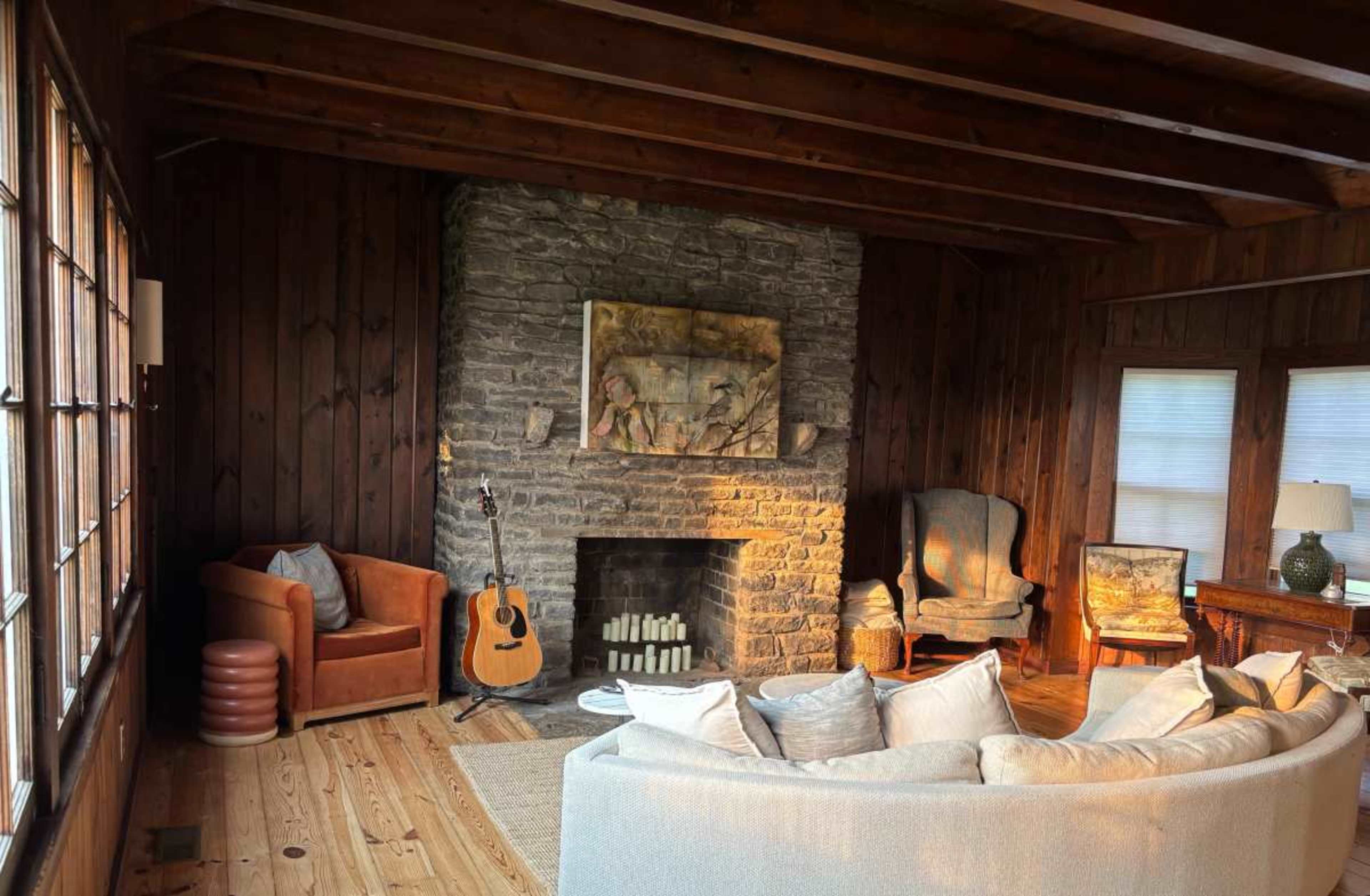 The image shows a cozy living room featuring a stone fireplace, wooden walls, a large sofa, an armchair, and a guitar leaning against the fireplace.