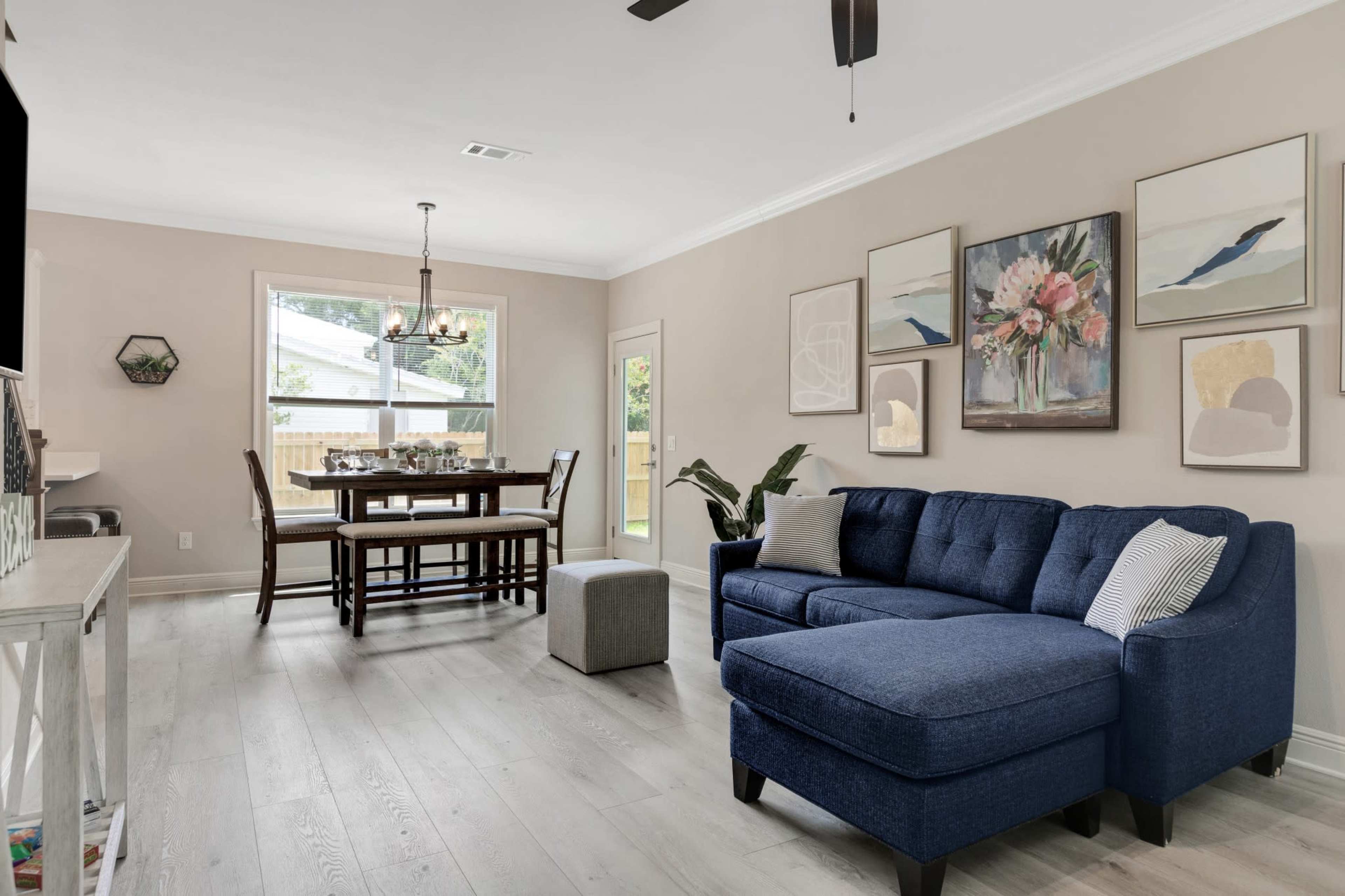 A cozy living room features a blue sectional sofa, a dining table set for six, and wall art in a neutral-toned space with natural light coming from a window.