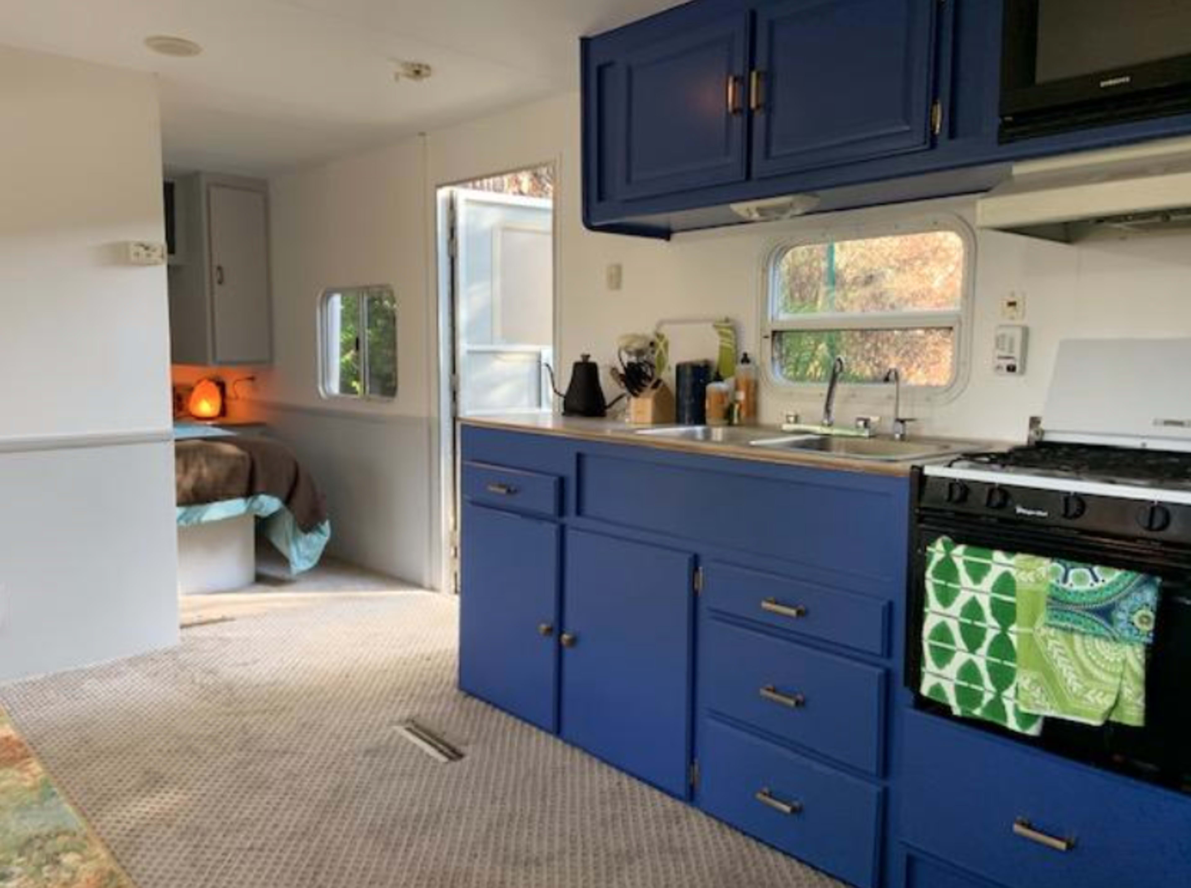 The image shows a small kitchen area with blue cabinets, a stove, and a sink, leading to a cozy bedroom adjacent to it.