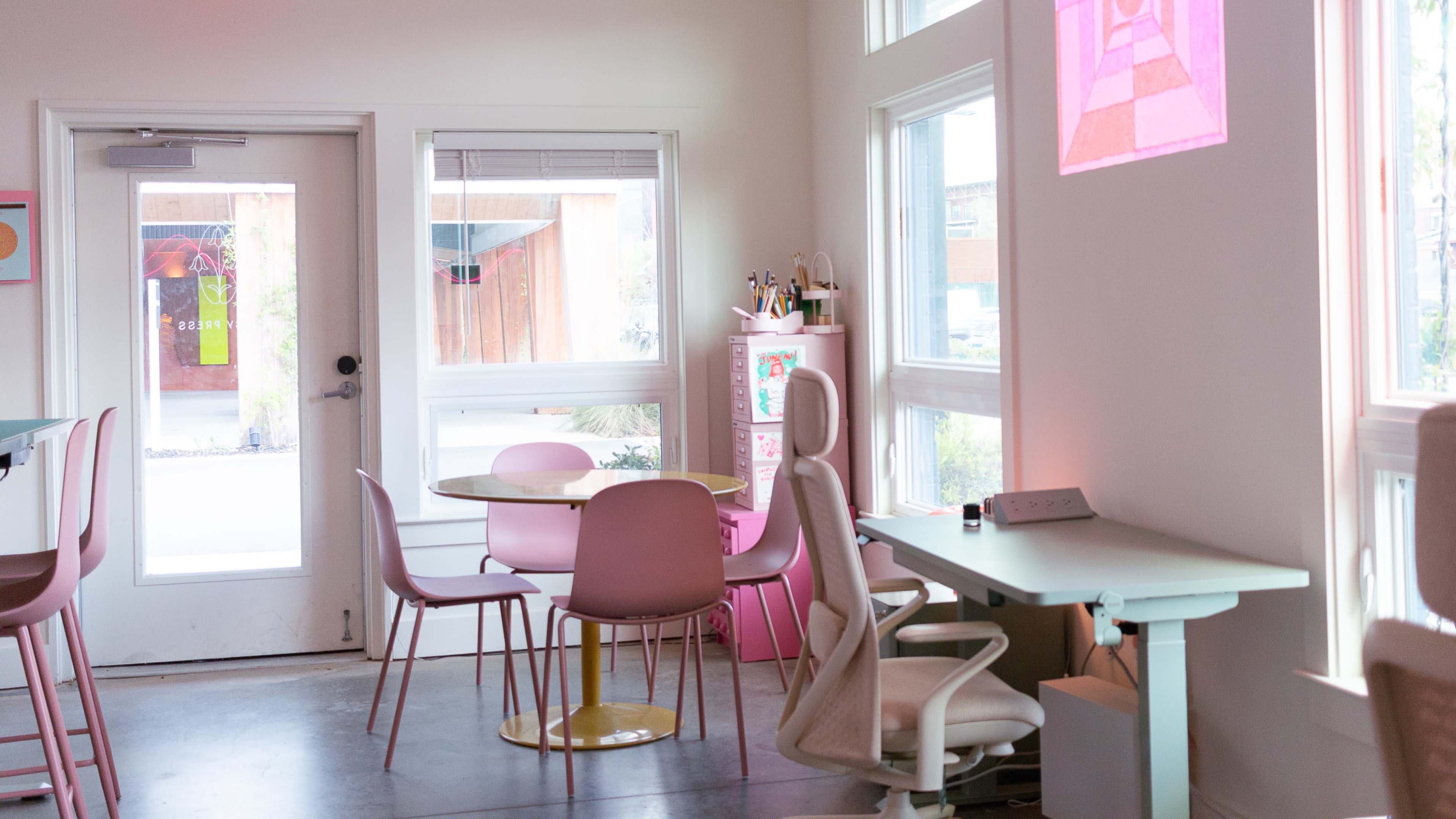 The image shows a modern cafe interior with pastel-colored furniture, including a round table, pink chairs, and a desk by the window.