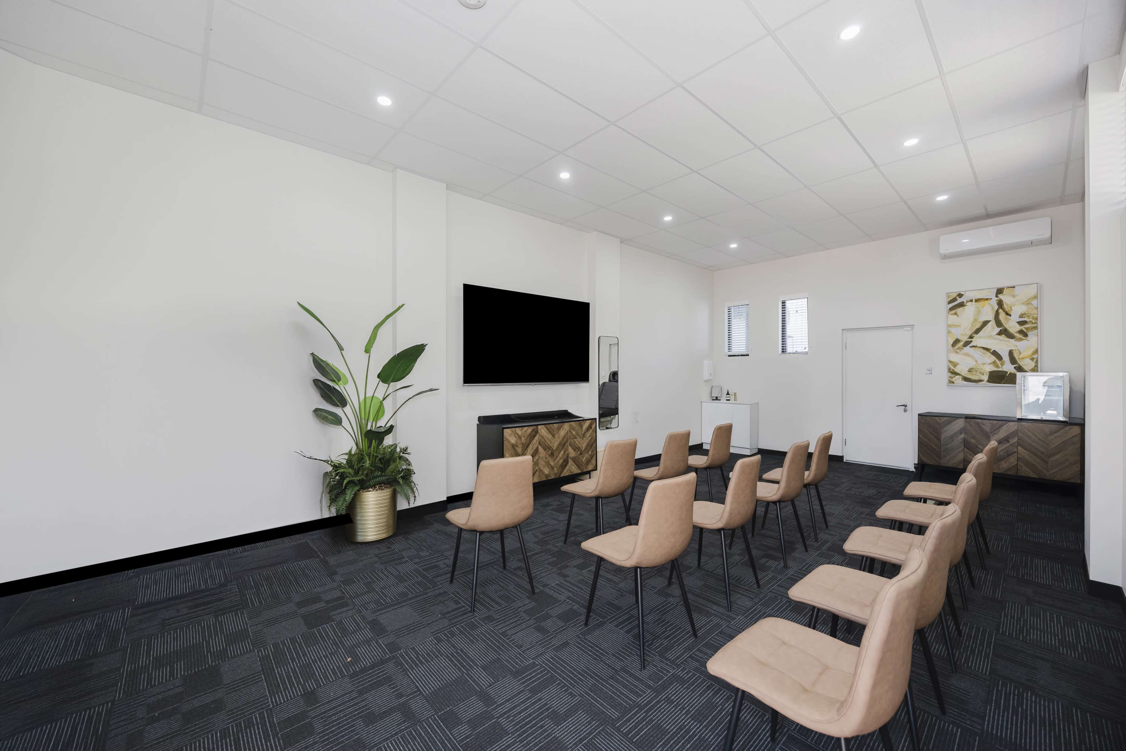 The image shows a modern meeting room with several chairs arranged in rows facing a wall-mounted television and a small plant in a corner.