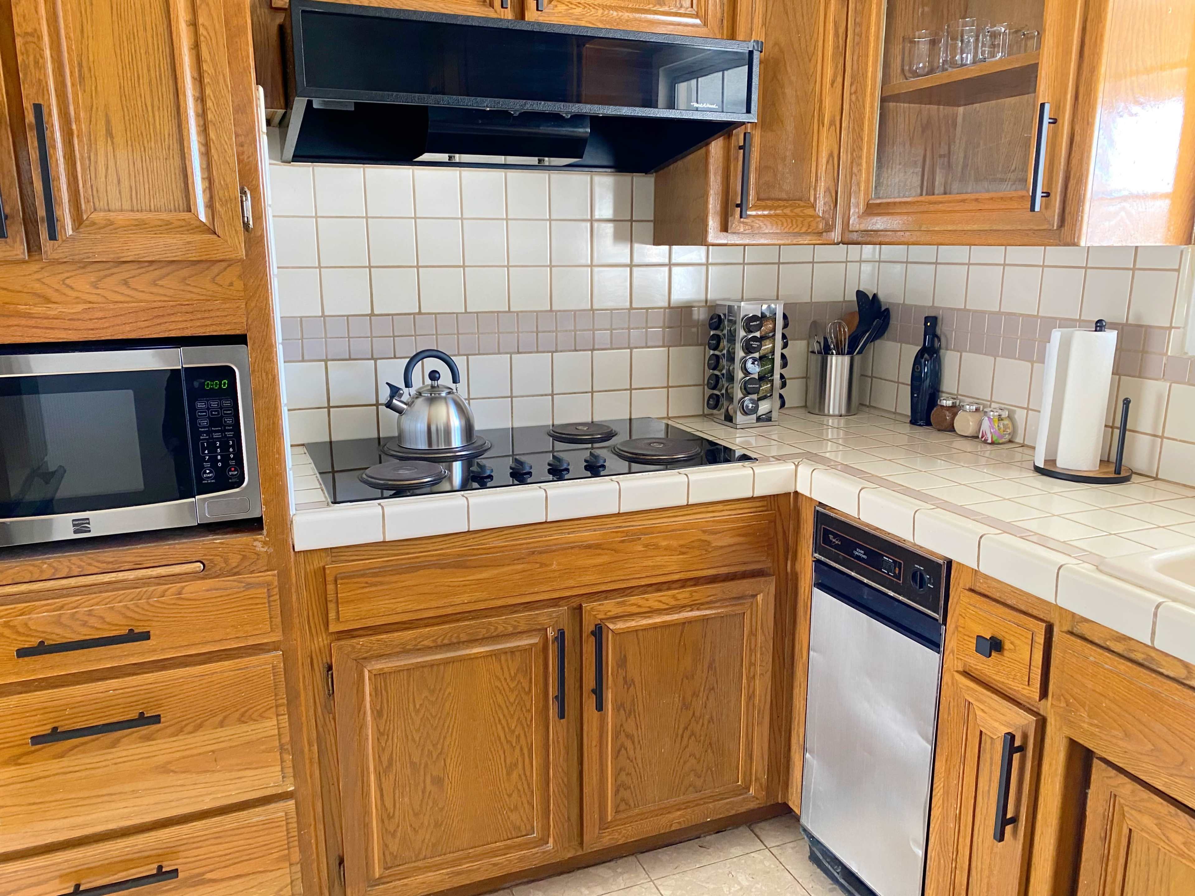 The kitchen features wooden cabinets, a stainless steel dishwasher, a microwave, a kettle on the stove, and a countertop with tile and organized utensils.