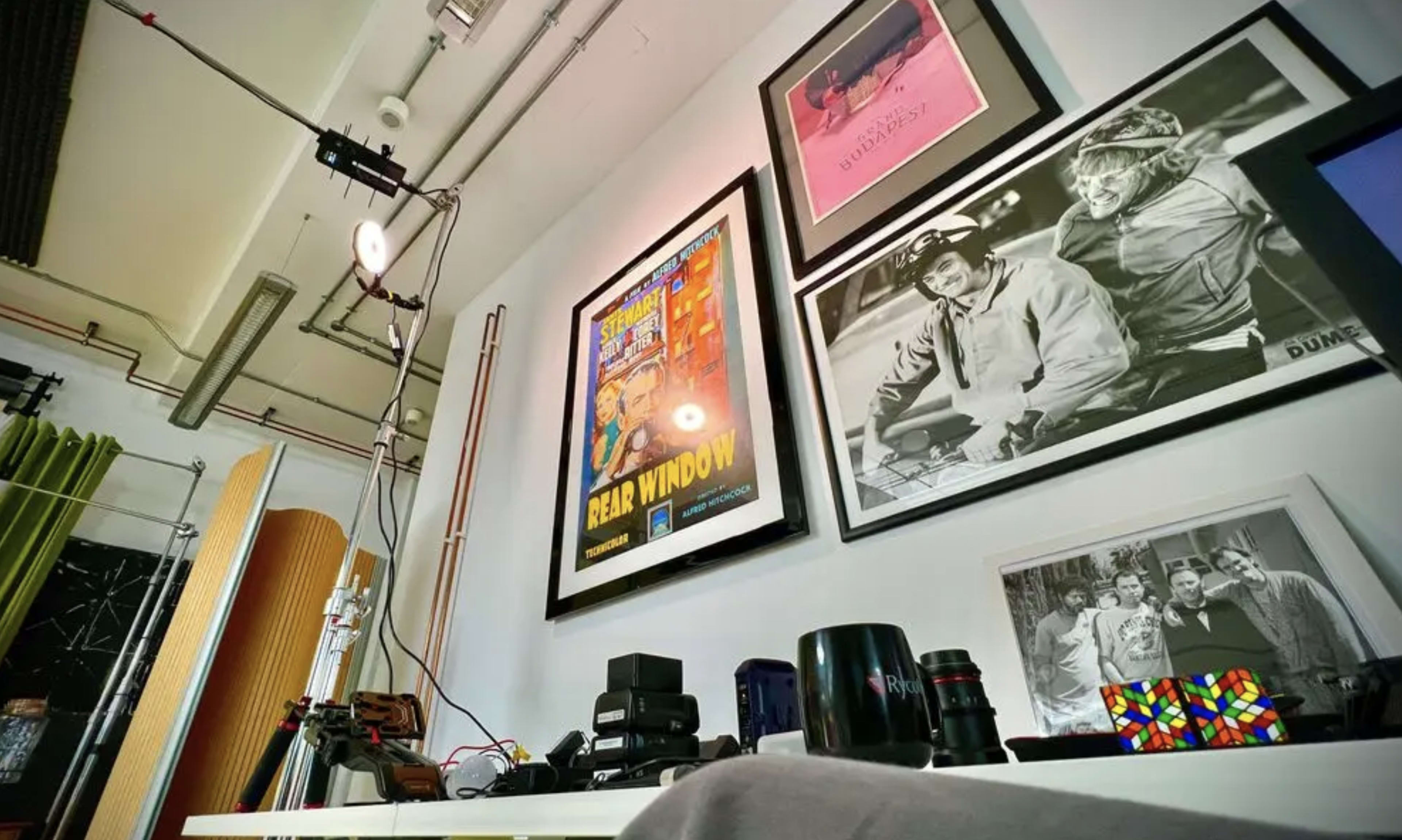 The image shows a corner of a room with various framed artworks and photographs displayed on the wall, along with a collection of objects on a flat surface.