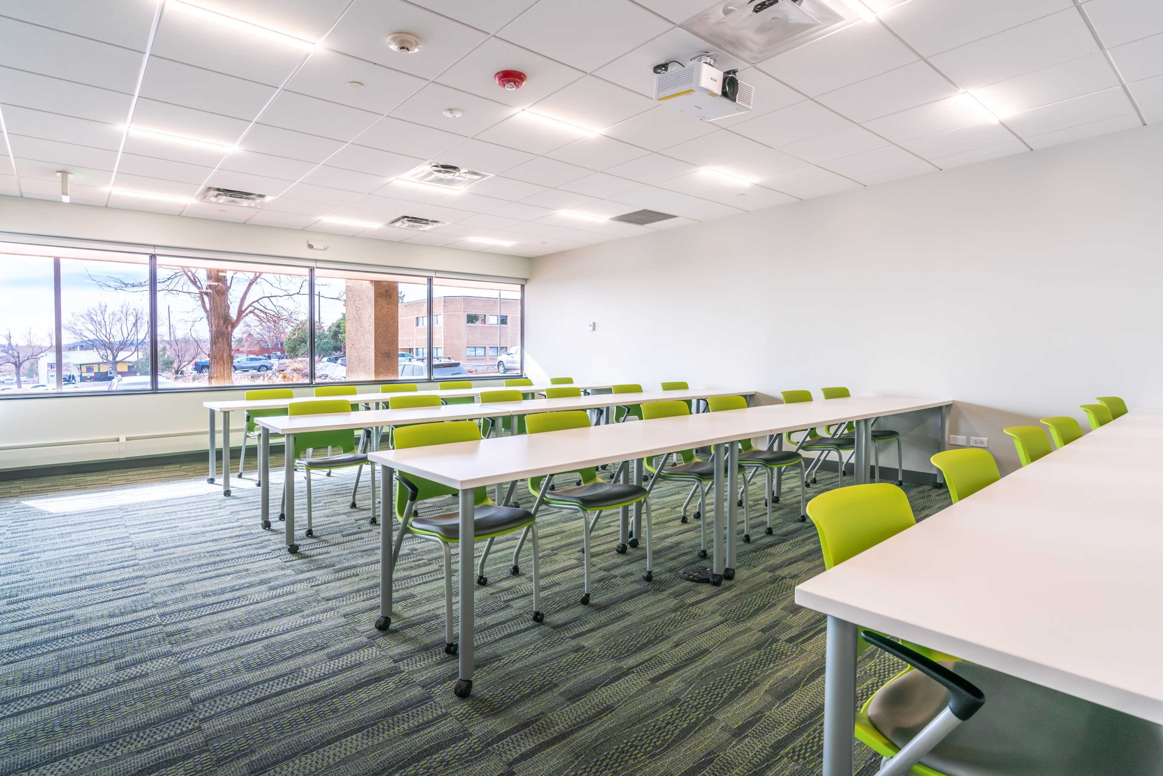 A modern classroom with rows of green chairs and white tables arranged neatly, illuminated by natural light from large windows.