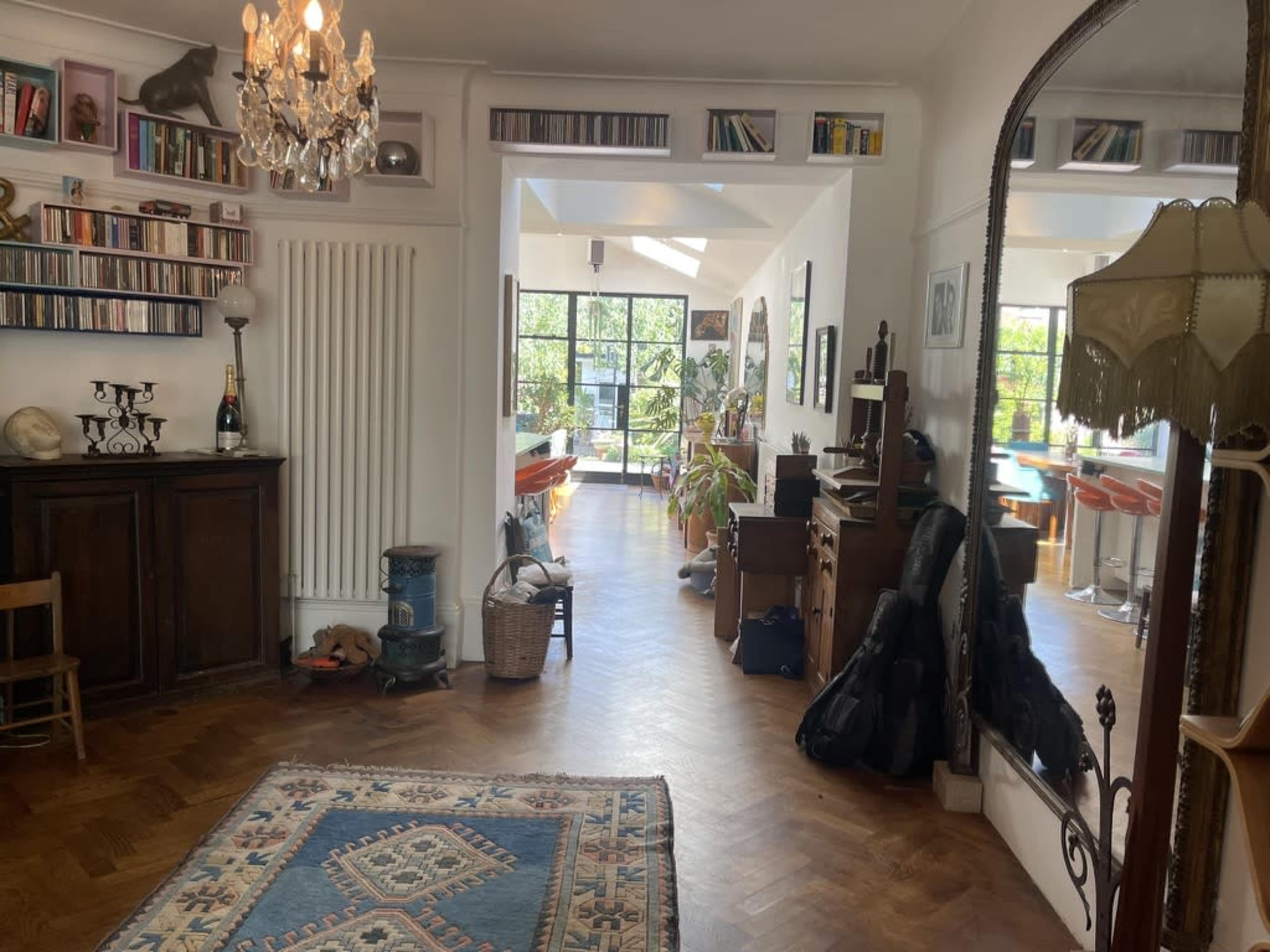 The image shows a bright, open living space with wooden flooring, a bookshelf filled with CDs, and a reflection of another room featuring orange furniture and large windows.