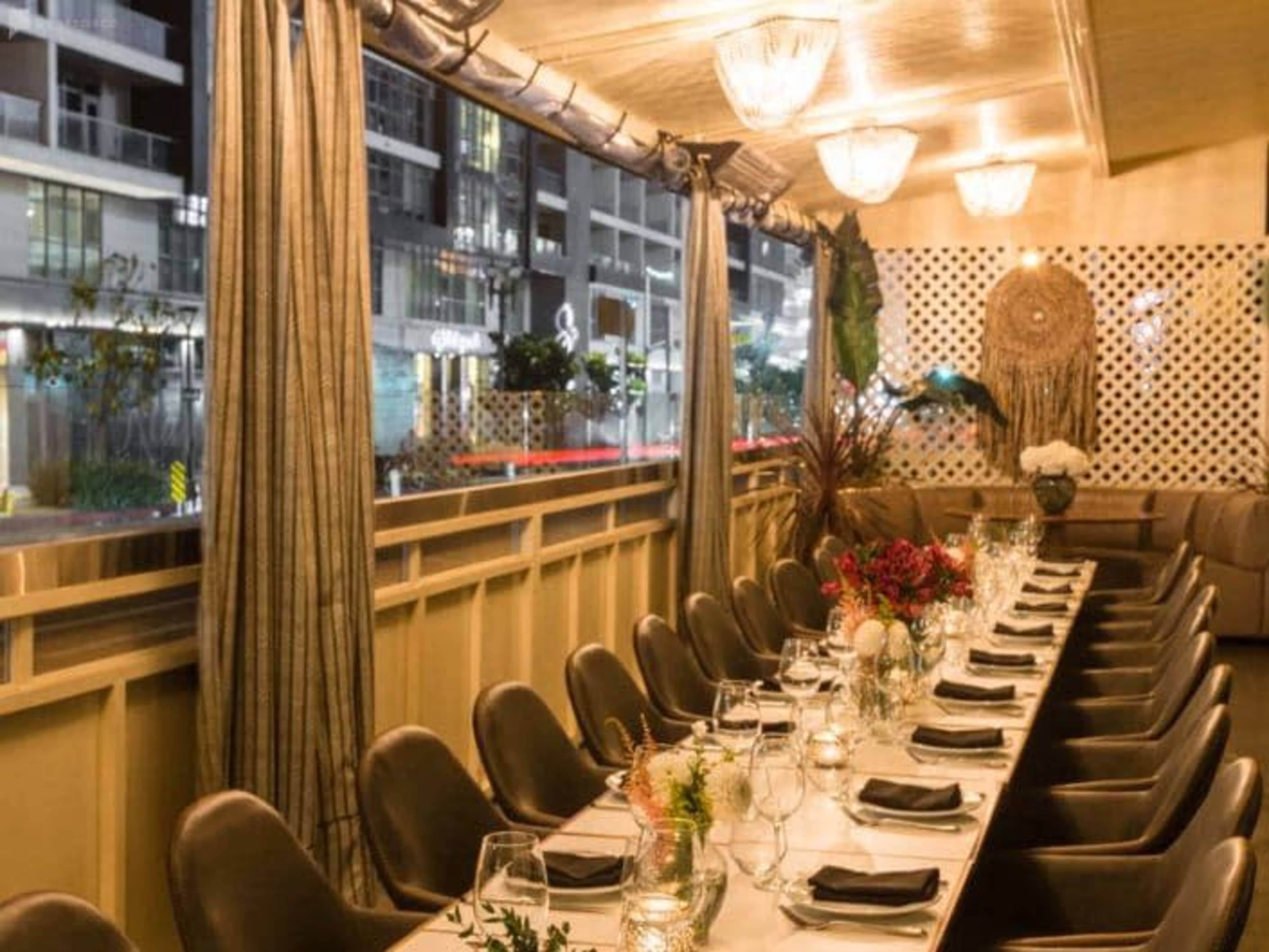 A long dining table is set with glasses and tableware in a brightly lit restaurant with large windows.