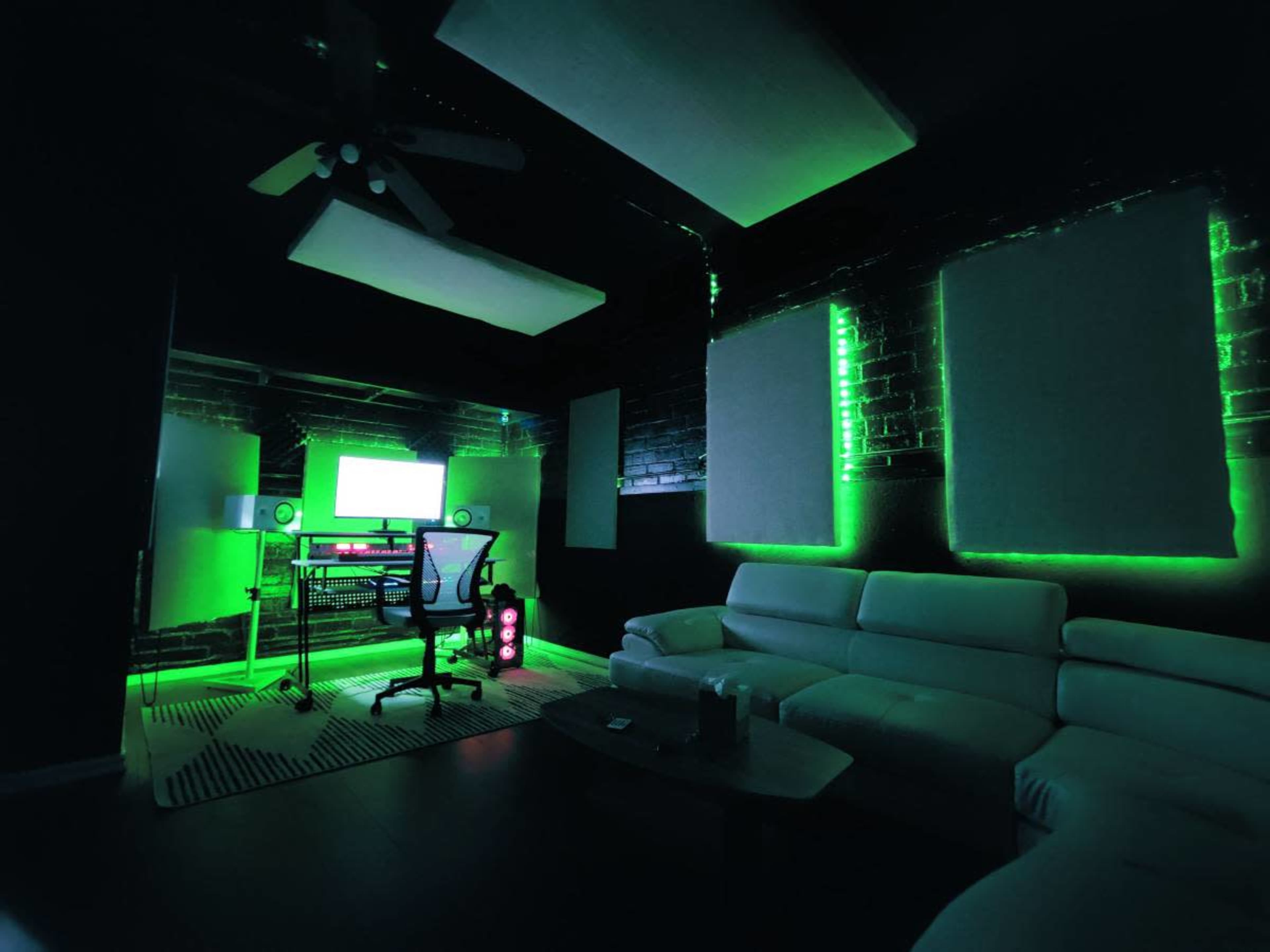 A modern studio workspace features a white leather couch, a sleek desk with dual monitors, and green ambient lighting illuminating the walls.