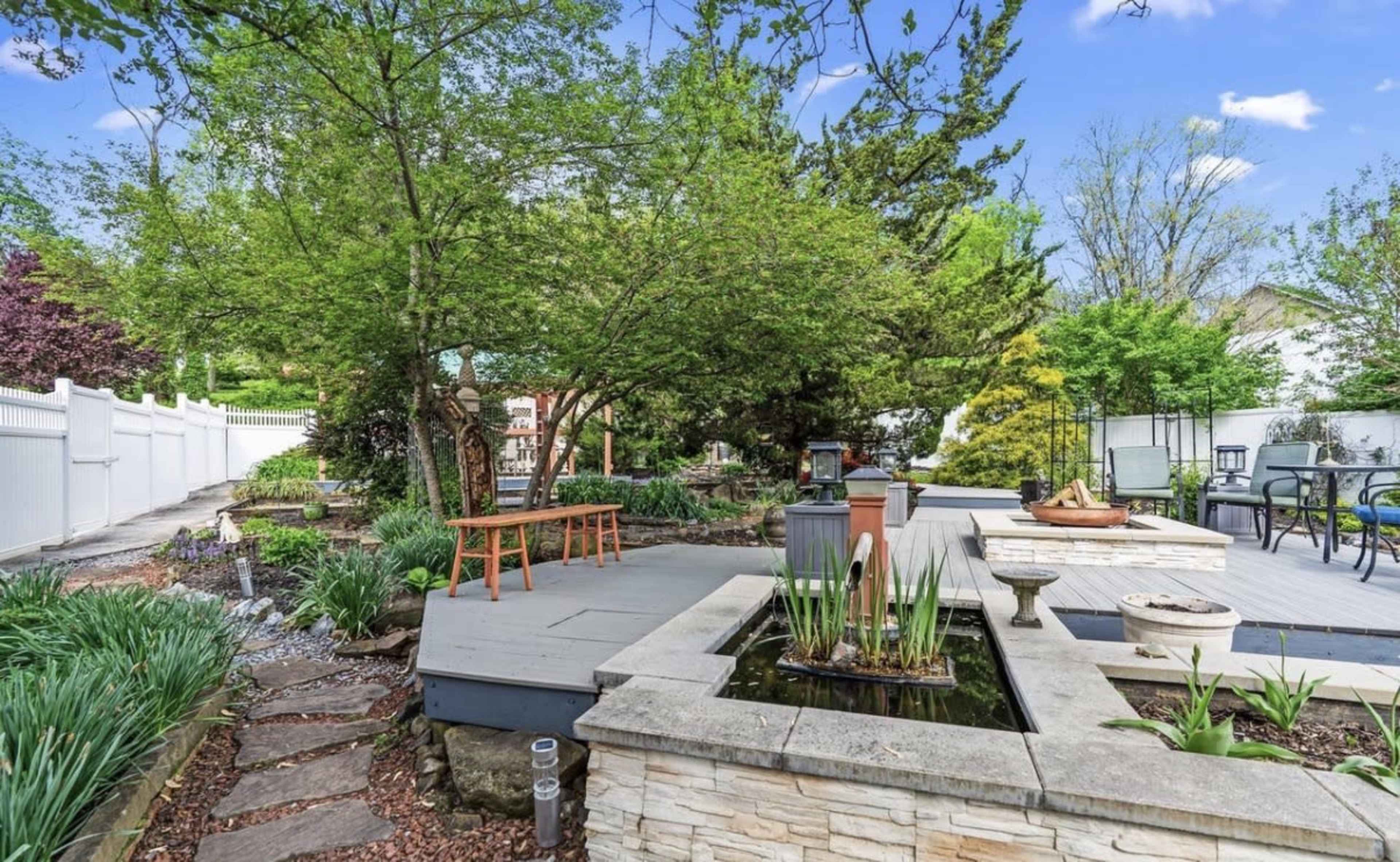 A serene backyard features a wooden bench beside a pond, surrounded by greenery and stone pathways, with a white fence in the background.