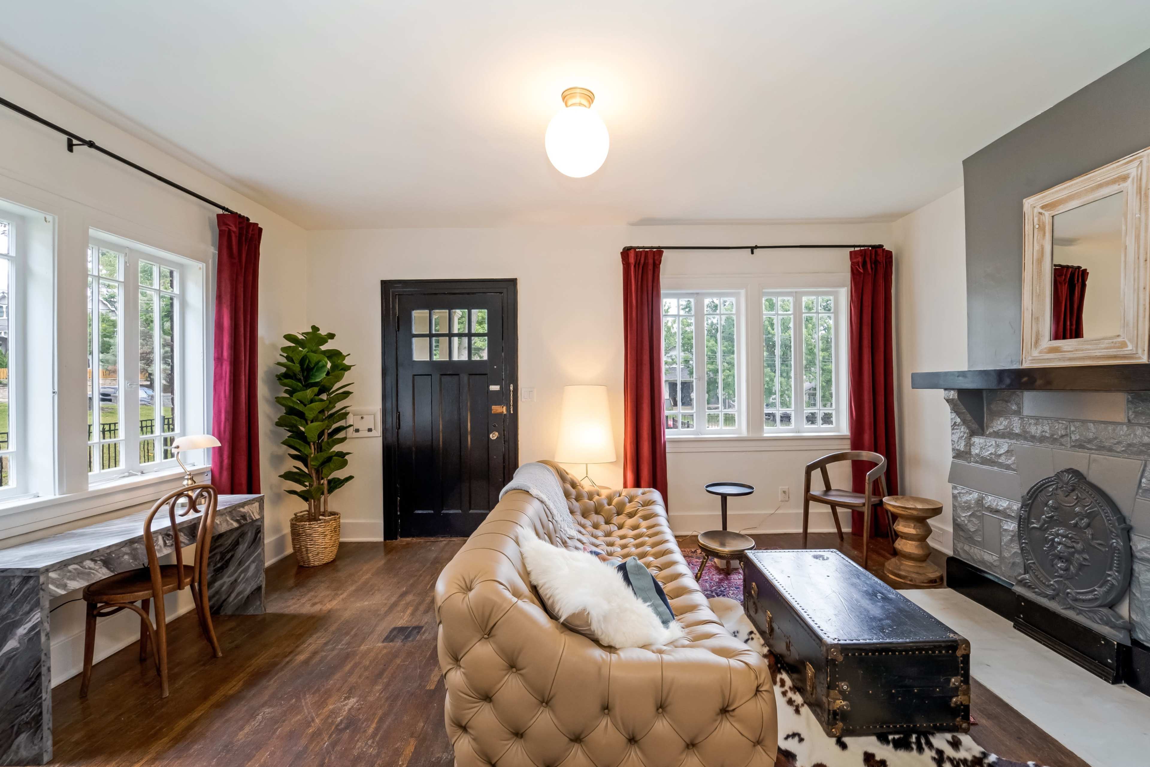 The image shows a living room with a tufted sofa, a black front door, and a decorative fireplace, featuring large windows and red curtains.