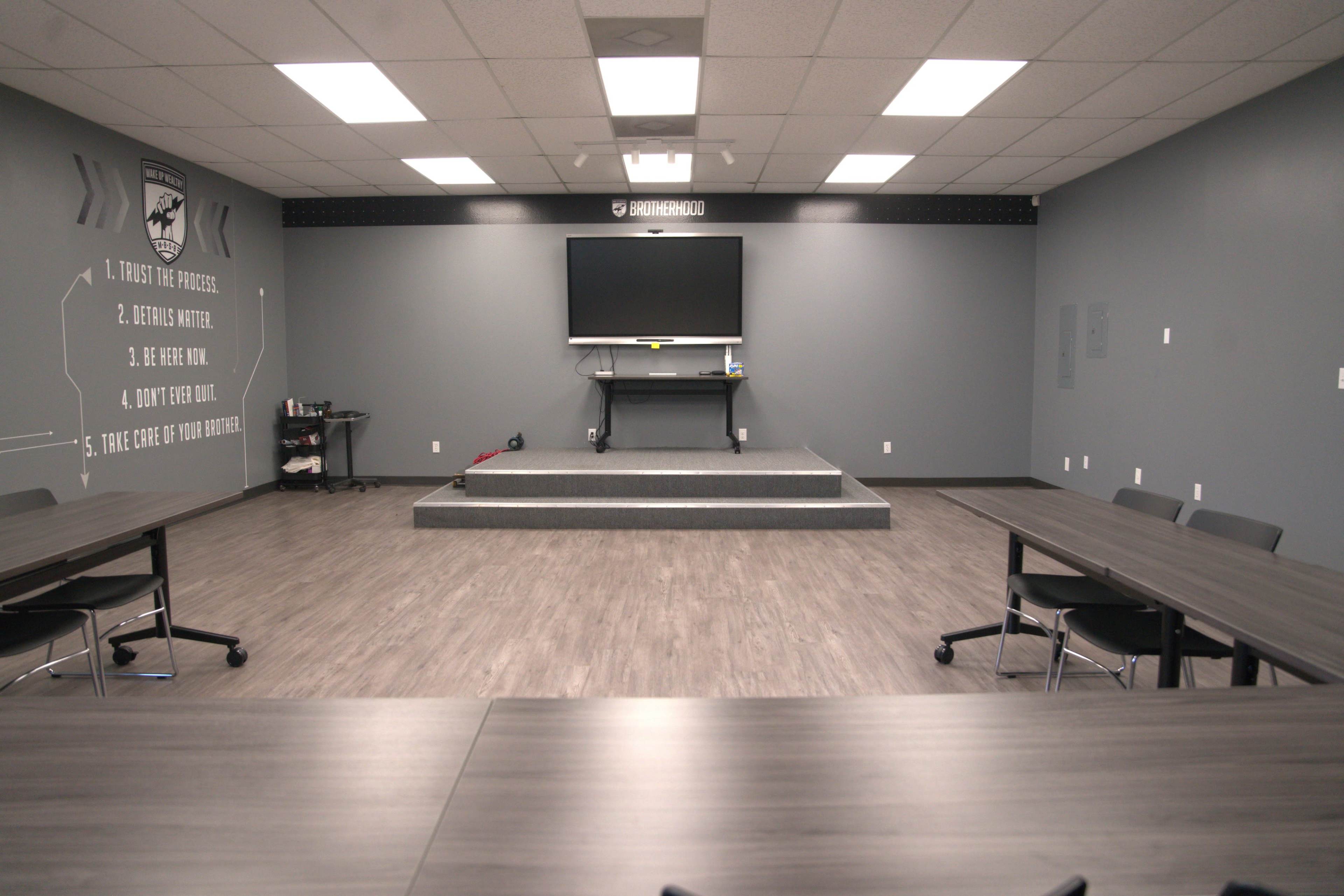 The image shows an empty training room with a stage, a television on the wall, and tables arranged in a U-shape facing the stage.