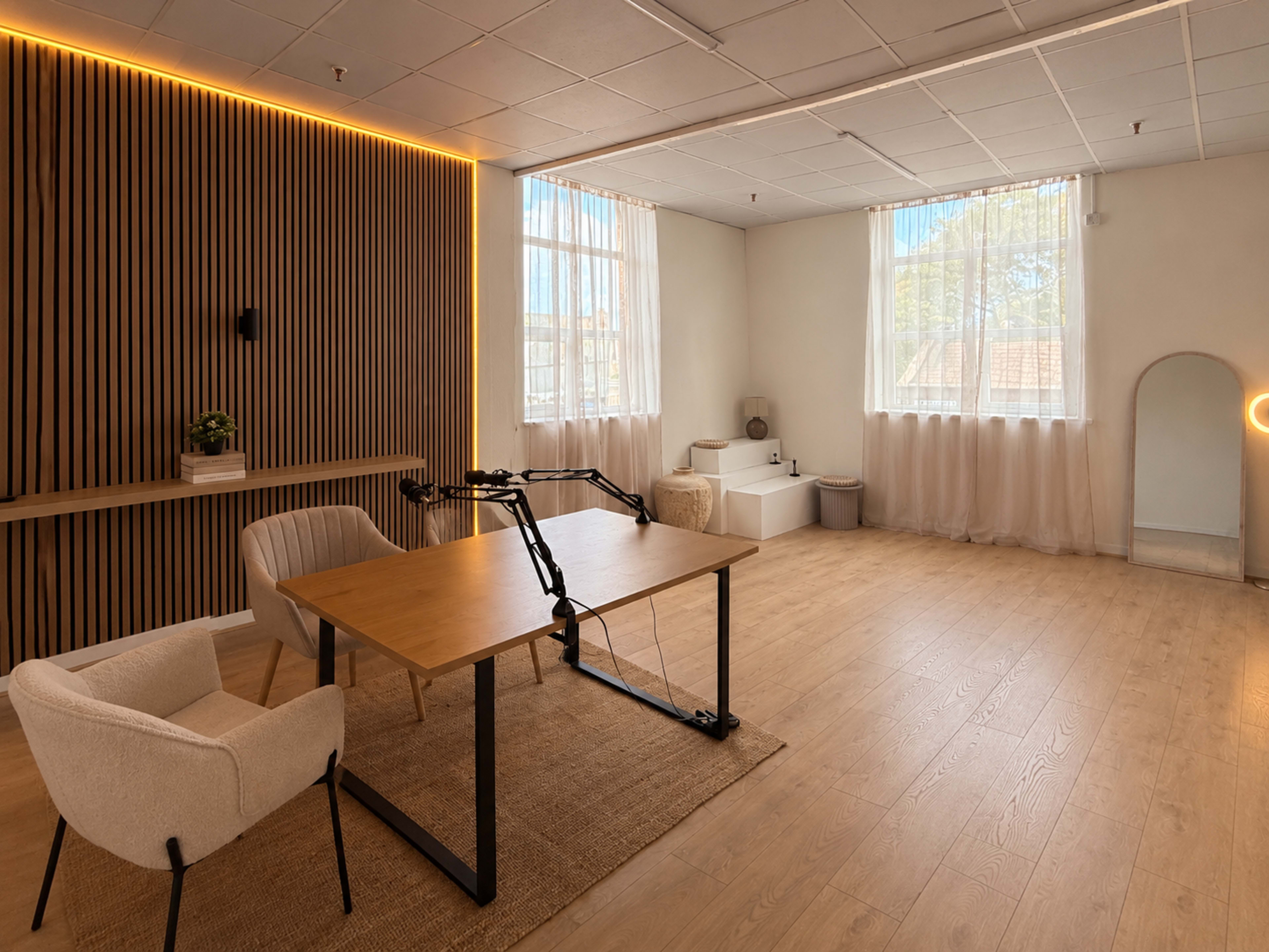 A bright, minimalist room features a wooden table with two chairs, large windows with sheer curtains, and a wall of vertical wood slats.