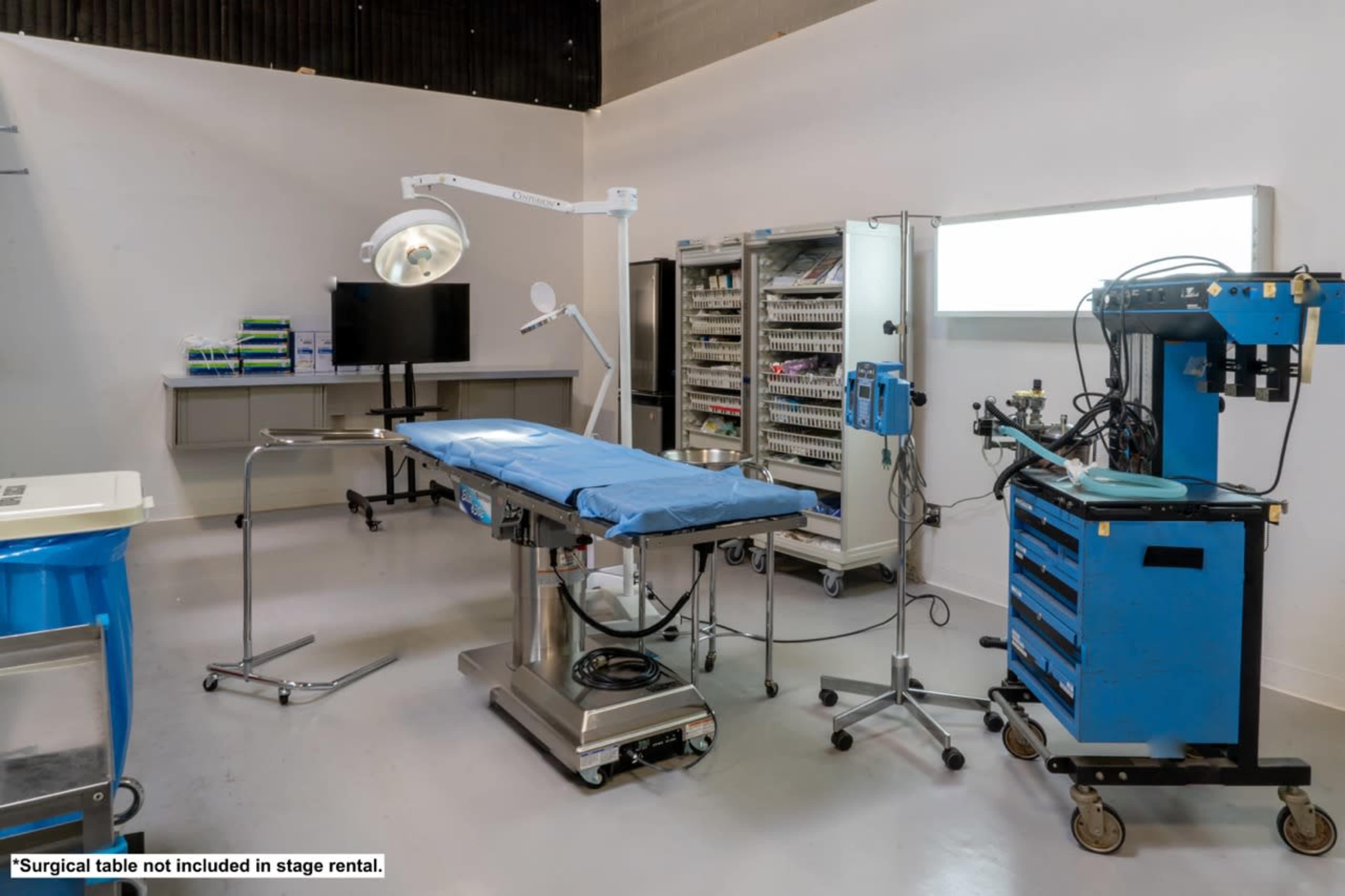 A well-equipped surgical room with a blue surgical table, medical instruments on a rolling cart, and an overhead surgical light.