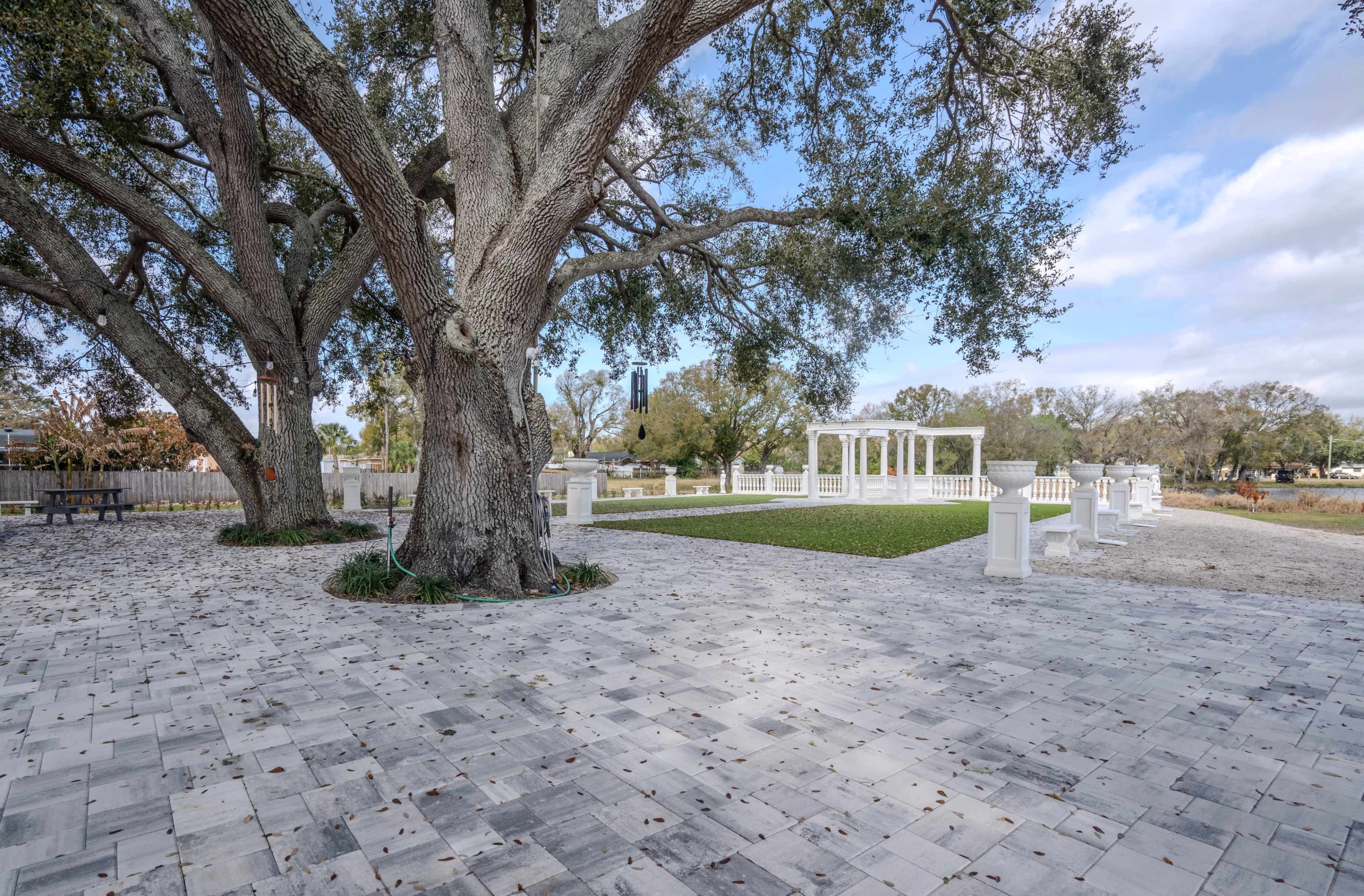The image shows a large tree in a landscaped area with a stone path, grass, and a white gazebo structure in the background.