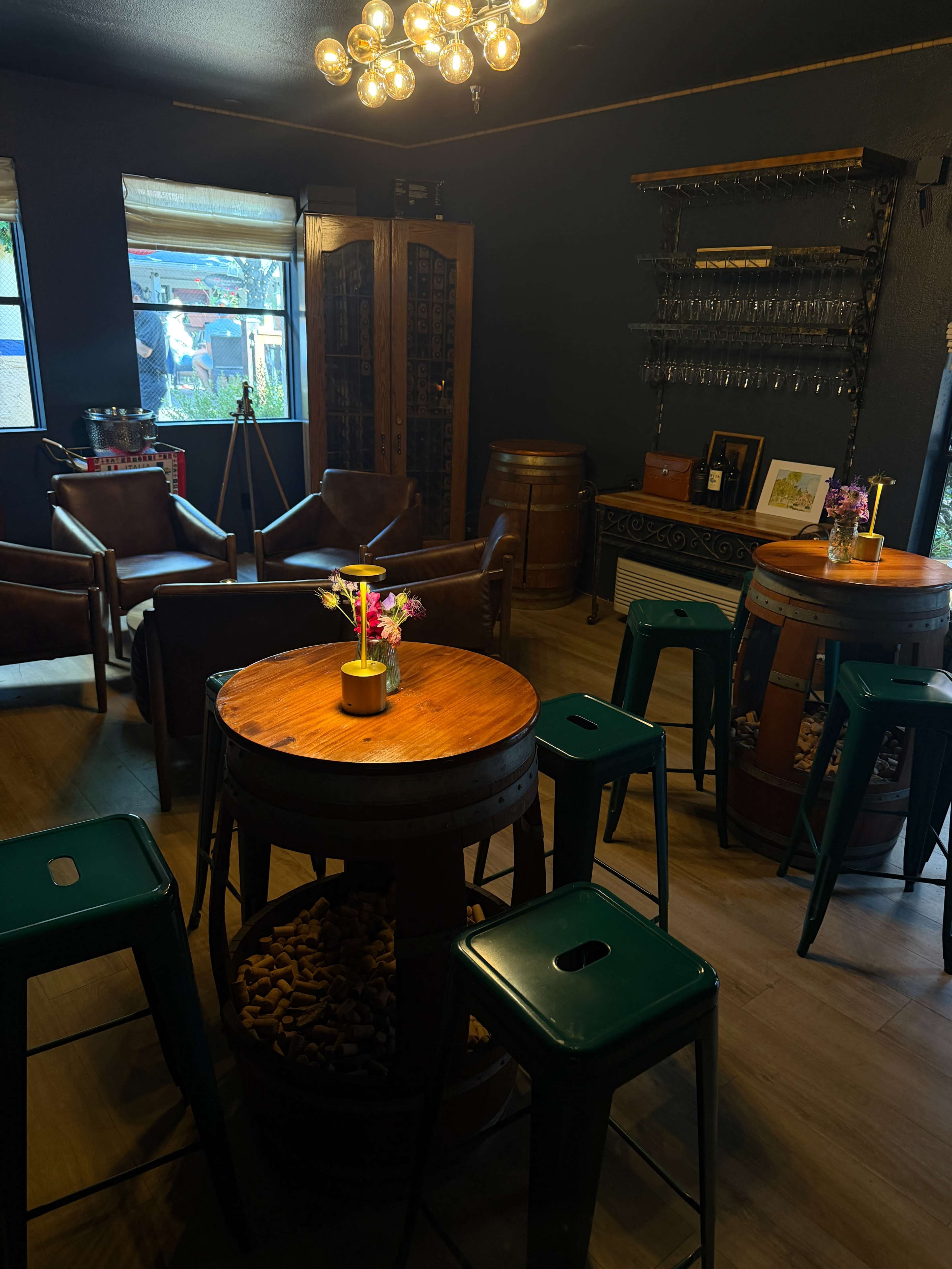 The image shows a cozy café interior featuring wooden tables, green metal stools, and a wall lined with wine glasses.