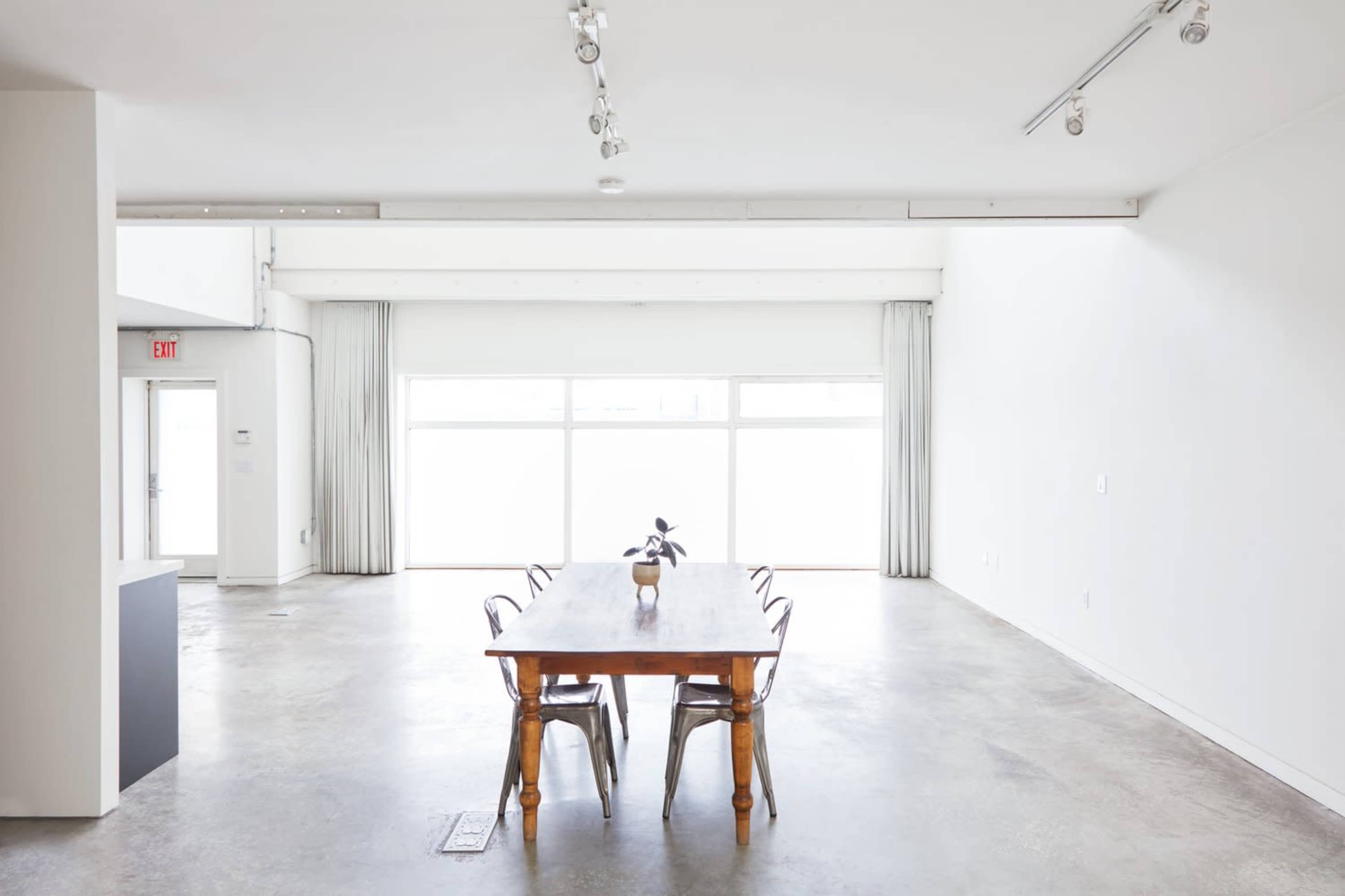 A wooden dining table with chairs is positioned in a spacious, well-lit room featuring large windows and minimal decor.
