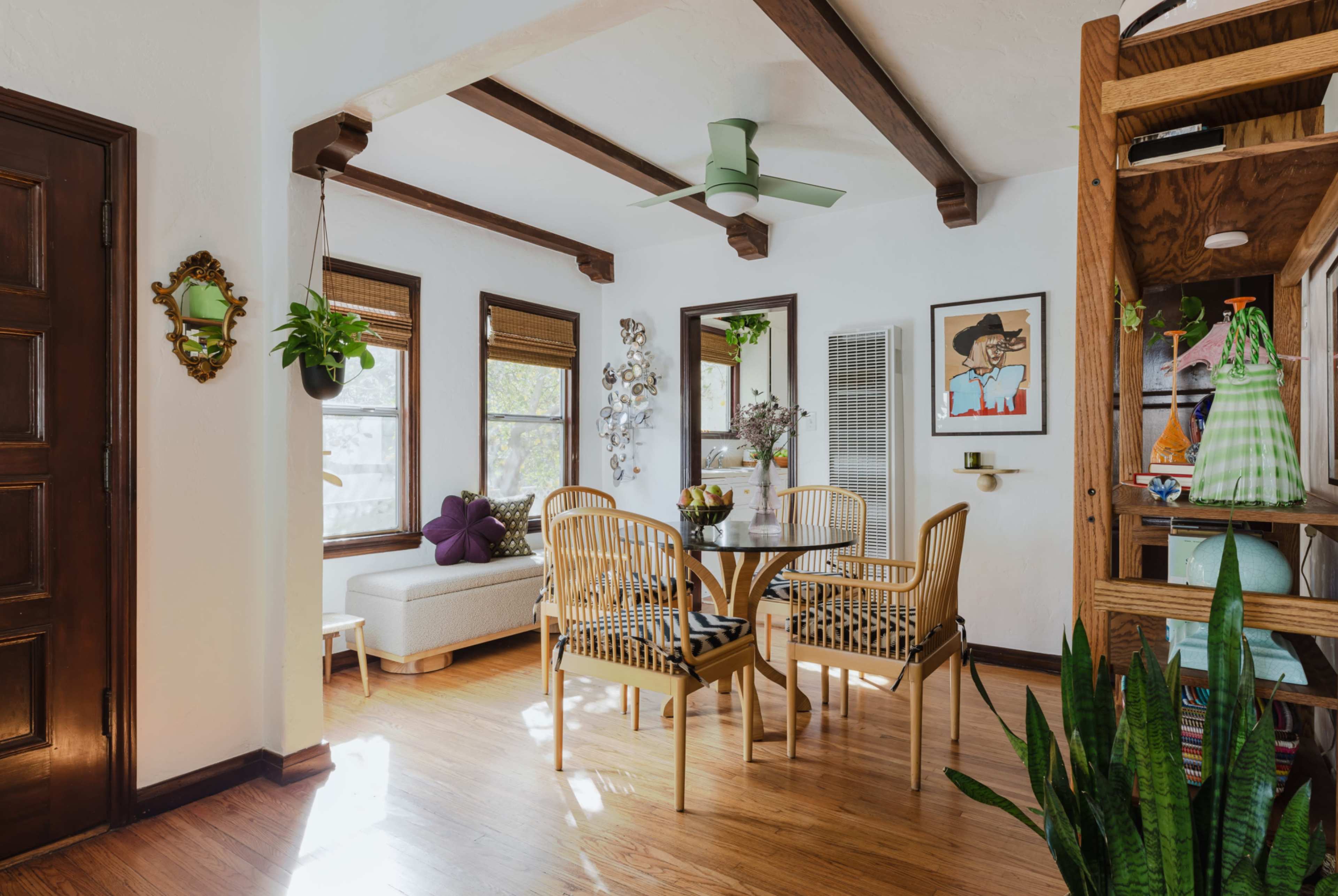 A dining area features a glass table surrounded by four chairs, with a sofa and plants near the windows, all set within a bright, open space.