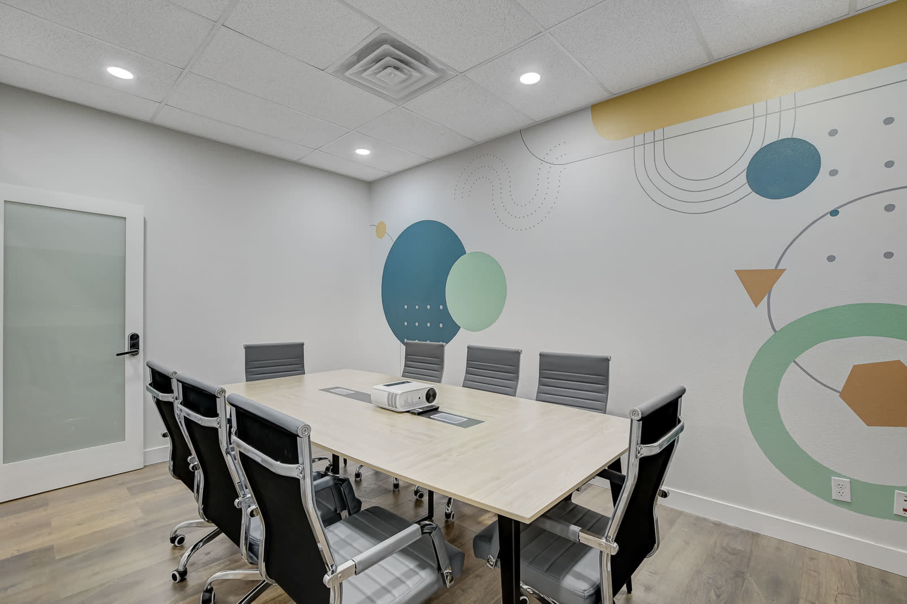 A conference room features a large table surrounded by chairs, with a projector on the table and colorful geometric designs on the wall.