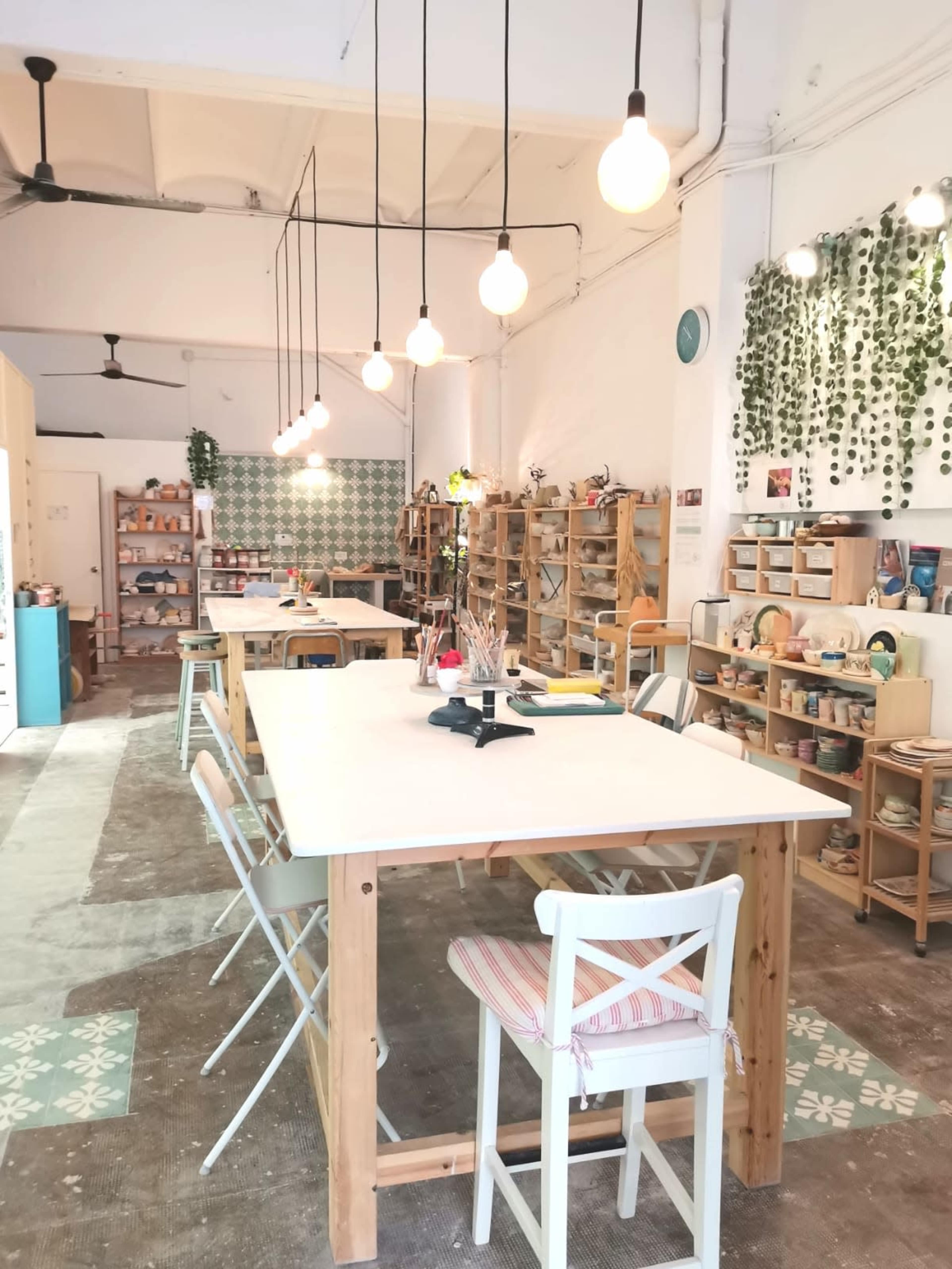 An airy craft studio with wooden tables, shelves filled with art supplies, and pendant lights hanging from the ceiling.