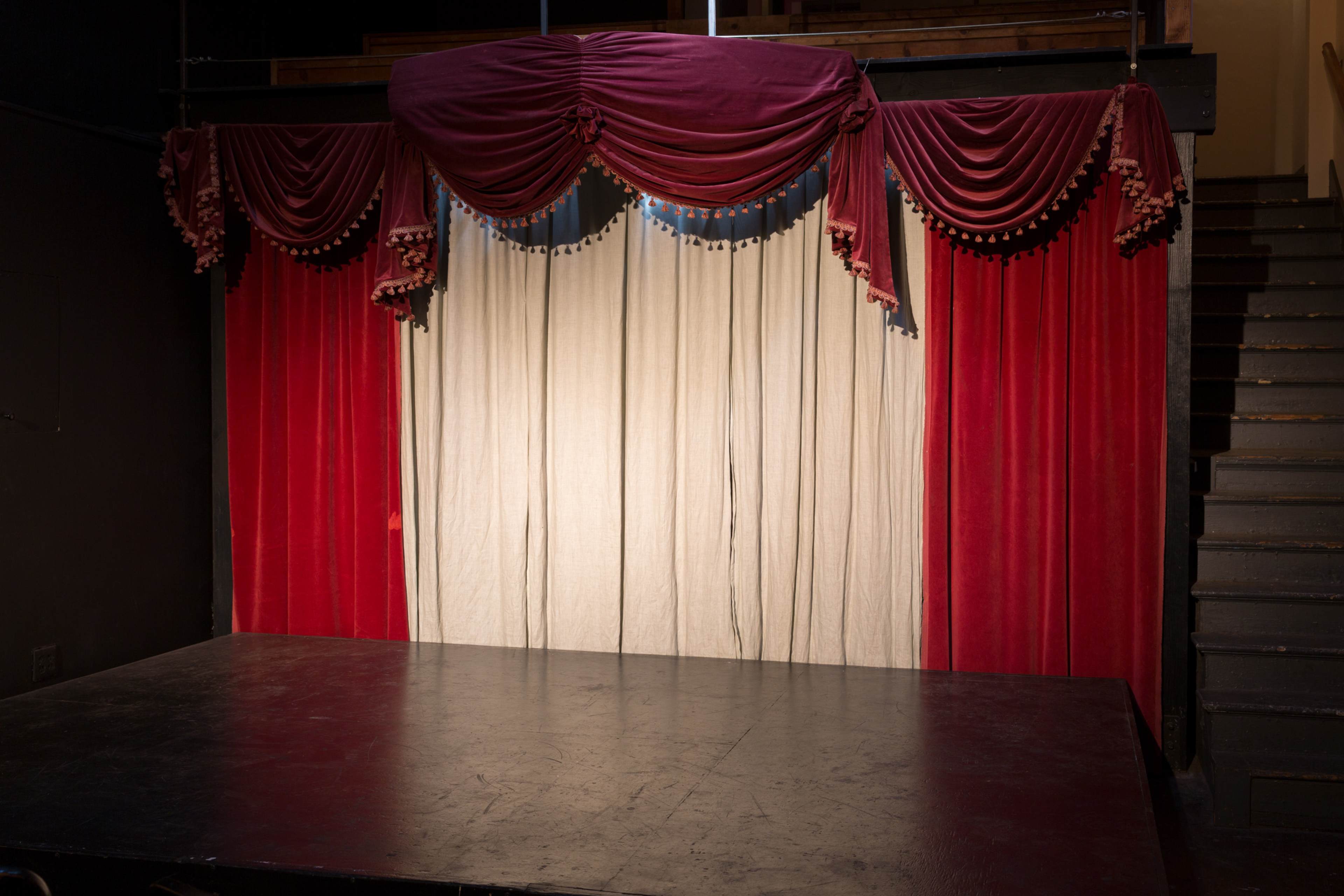 A stage with a velvet curtain in red and white, draped elegantly above a plain backdrop, framed by gathered fabric.