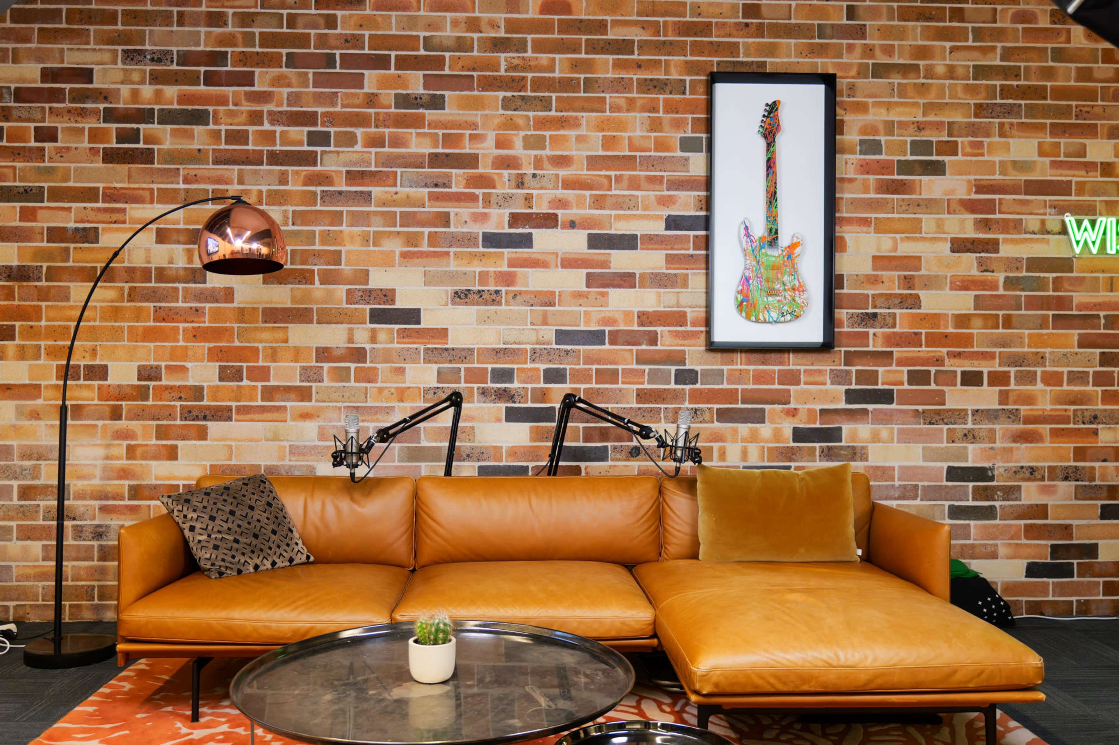 A brown leather sectional sofa is positioned against a brick wall, featuring a framed guitar artwork above and a modern floor lamp beside it.