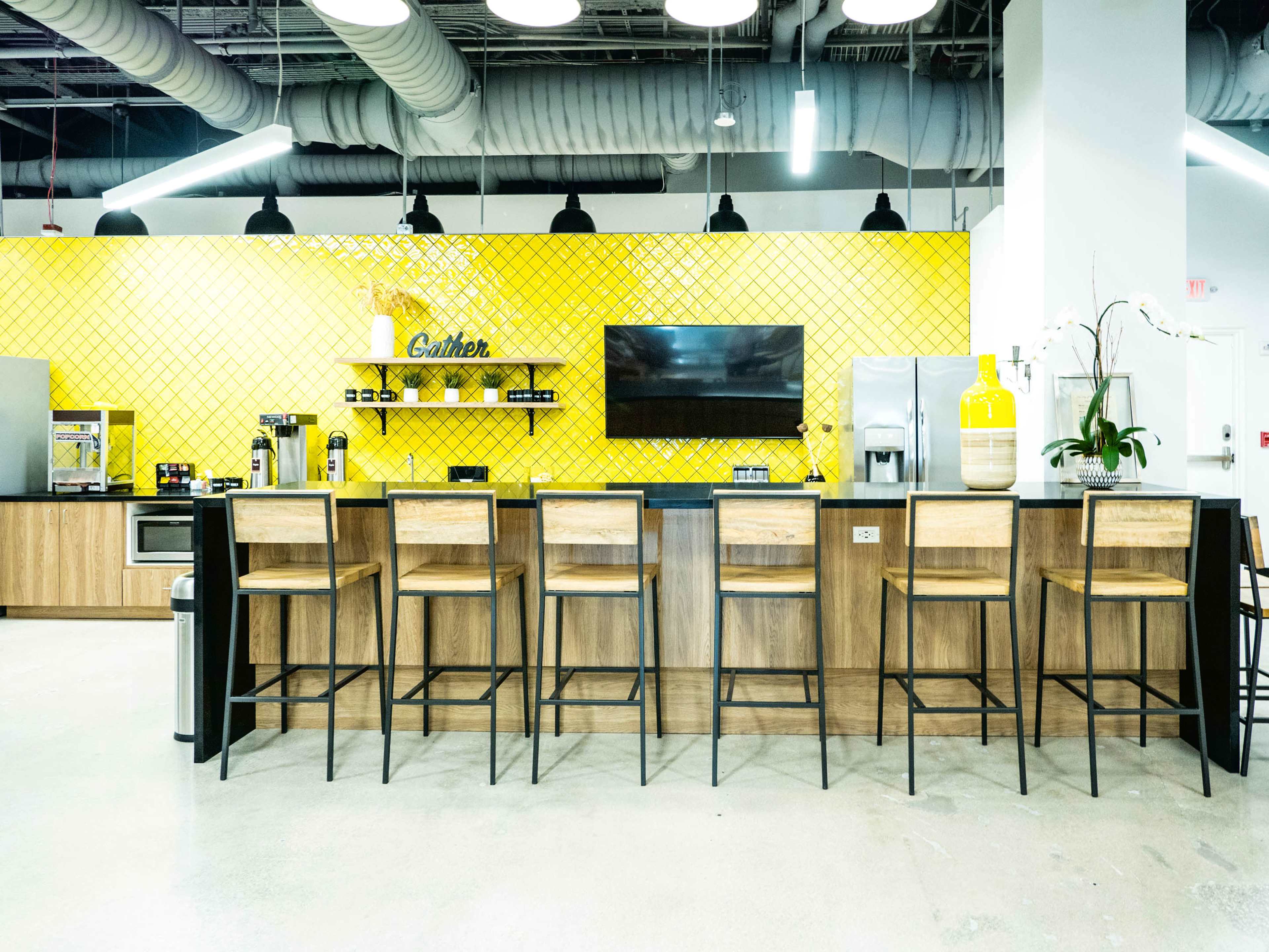 The image shows a modern kitchen with a yellow tiled wall, a large television, and a row of wooden bar stools in front of a countertop.