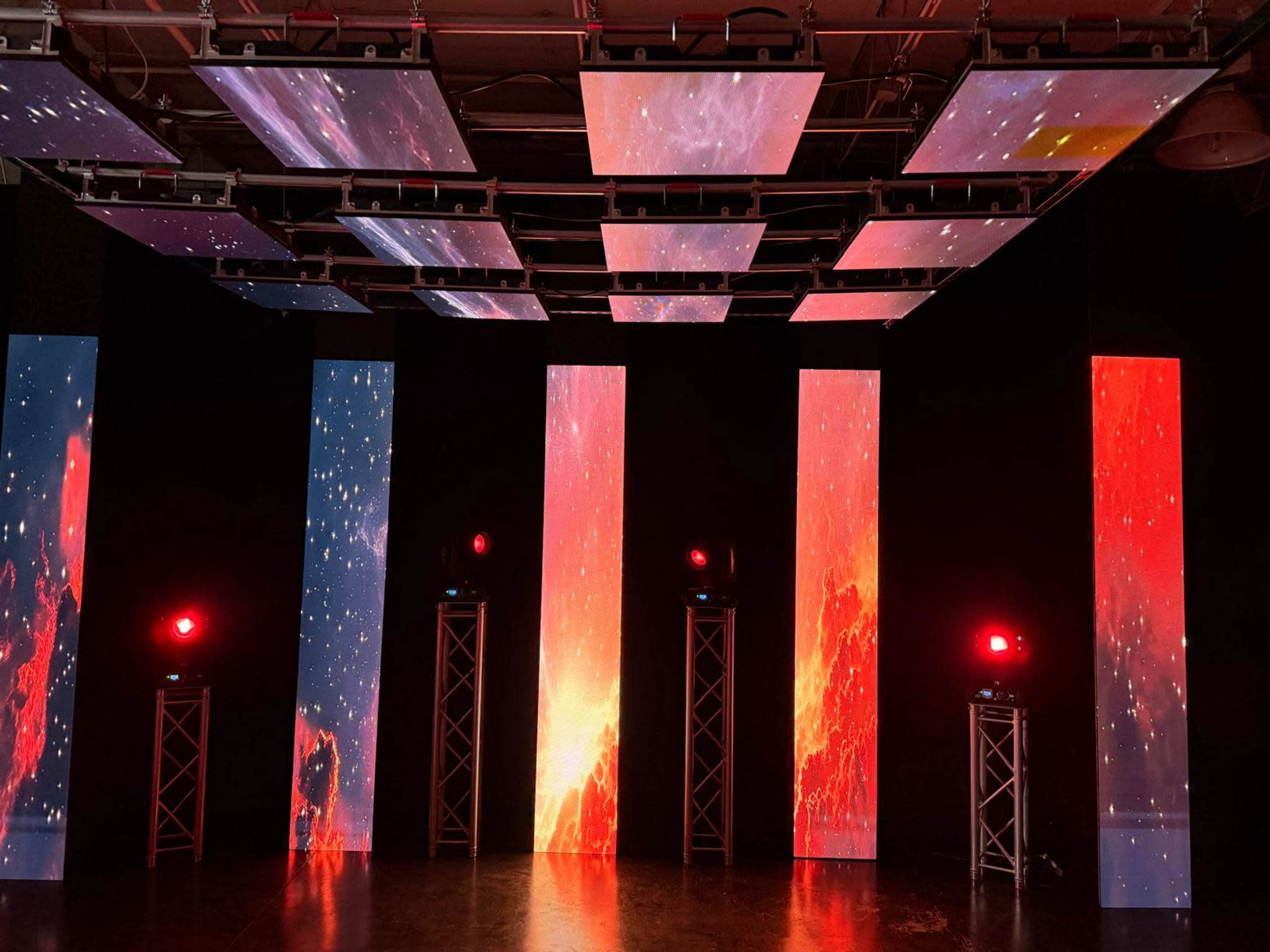 The scene features a dark room illuminated by multiple vertical video screens displaying vibrant cosmic imagery and red lights.