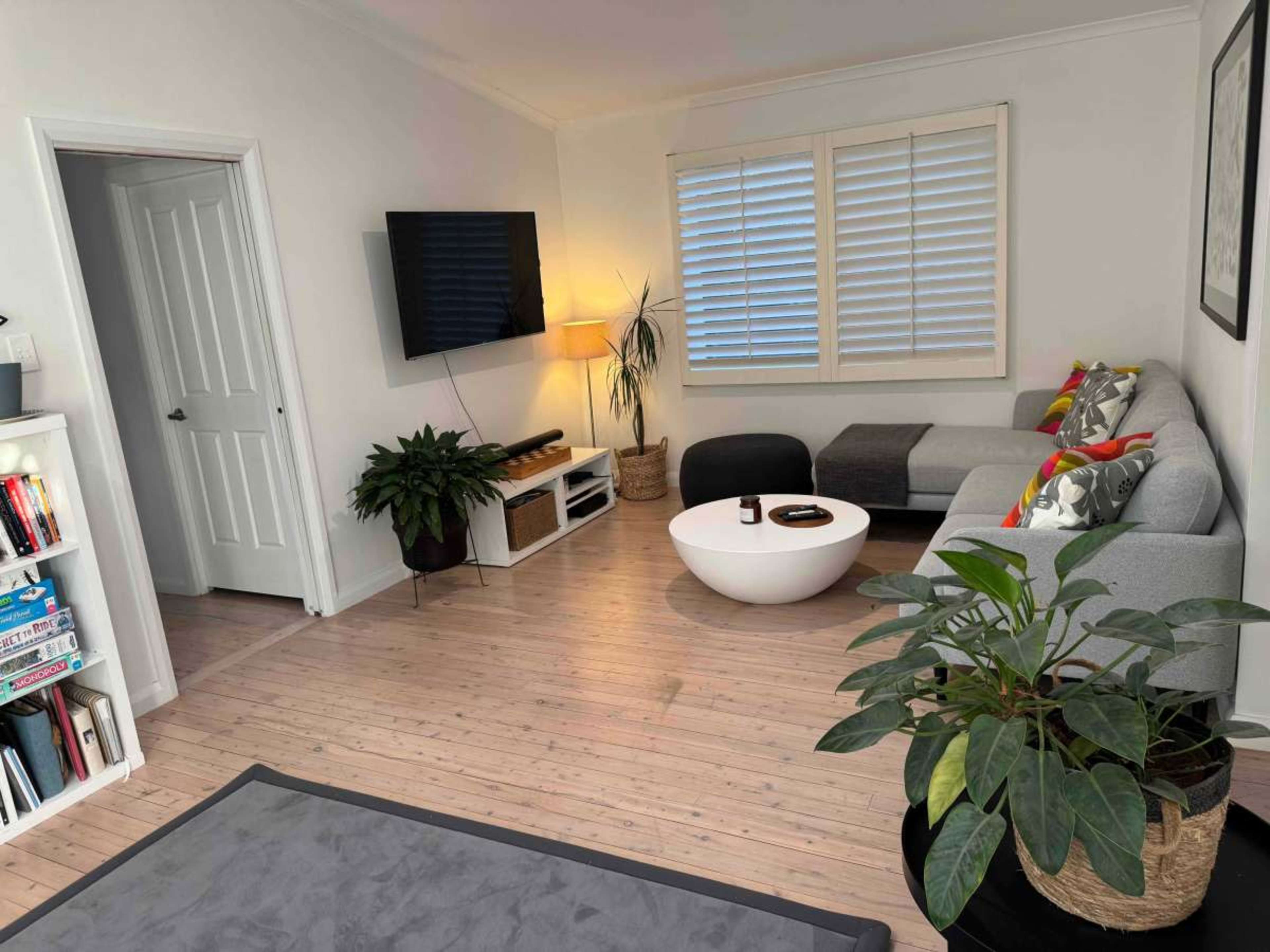 A modern living room features a gray sectional sofa, a round coffee table, a mounted television, and potted plants against light-colored walls and a wooden floor.